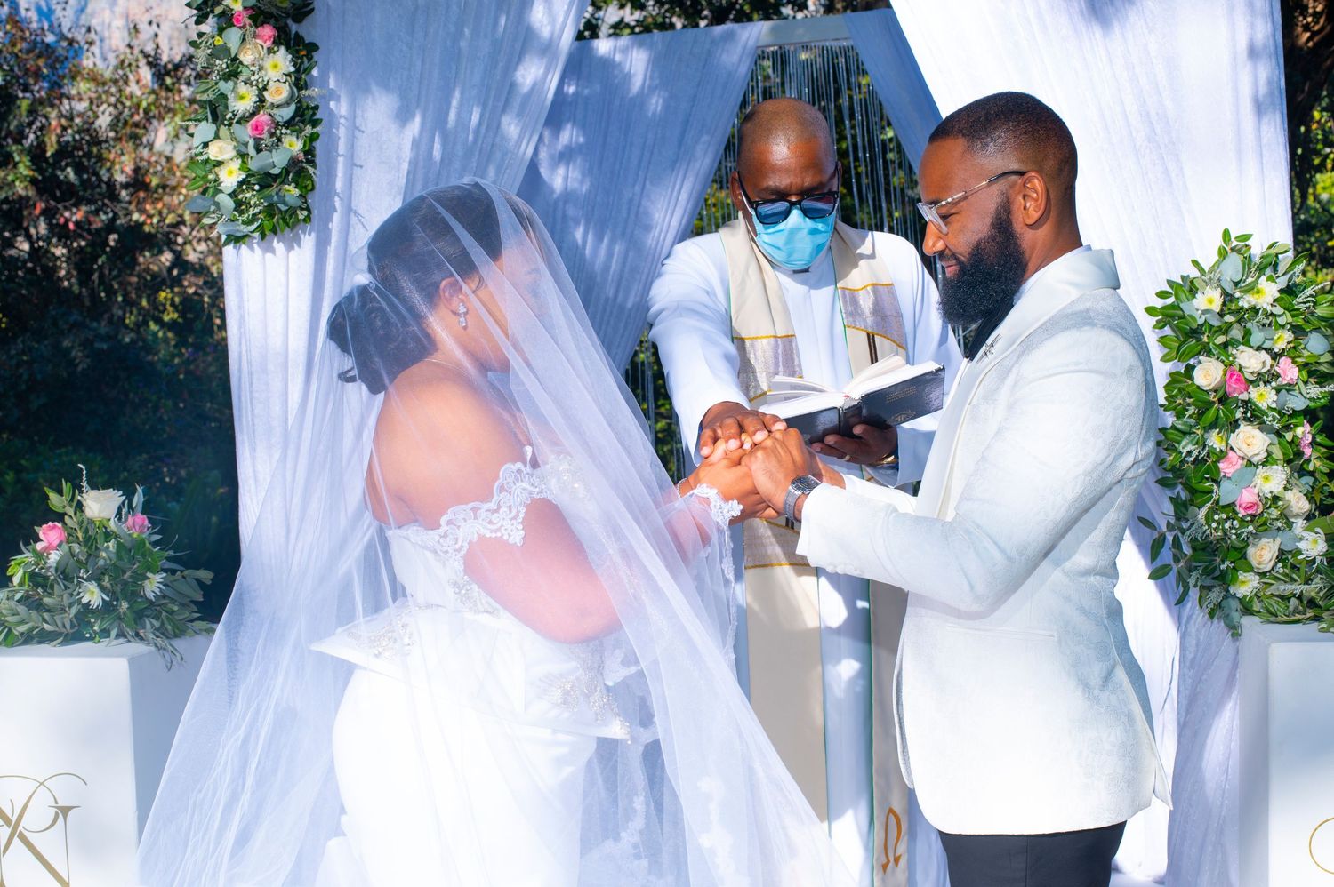 Bride and groom exchange rings at outdoor wedding ceremony with officiant wearing blue mask in background.