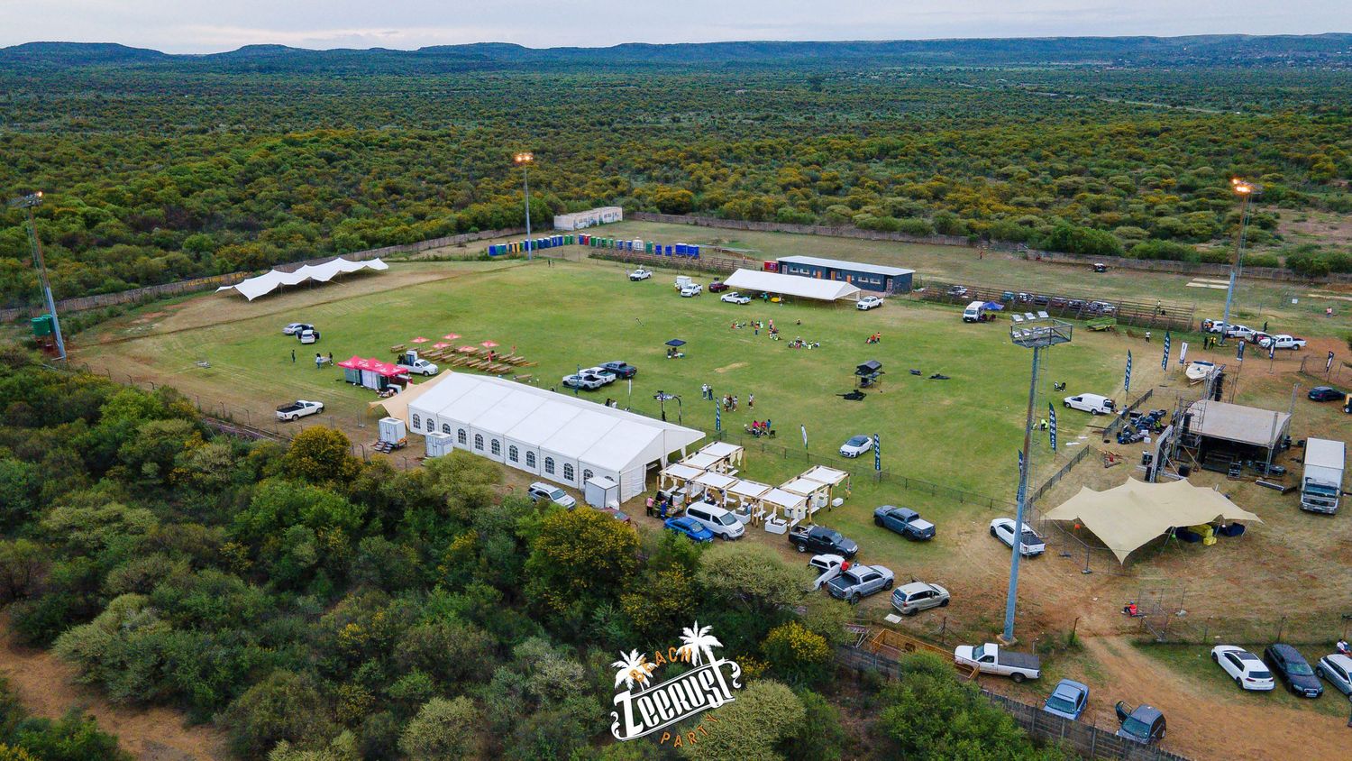 Aerial view of Zebonique outdoor event venue with green fields, white tents, parked cars, and surrounding bushveld landscape.