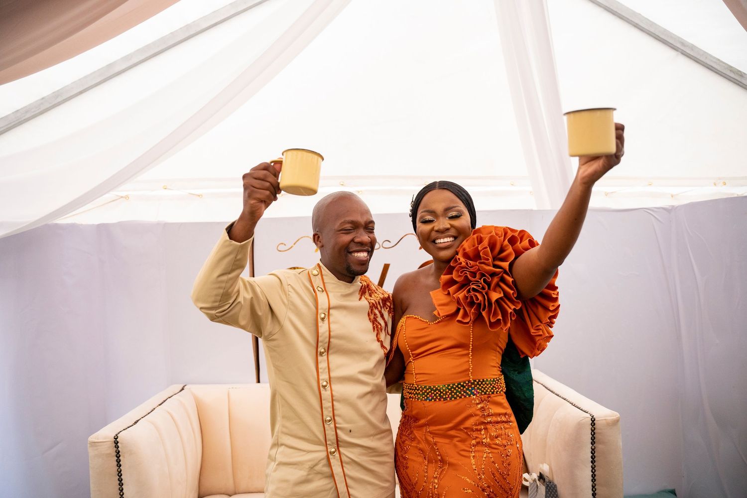 Joyful African couple in traditional orange and gold attire raise cups in celebratory toast at cultural wedding ceremony.