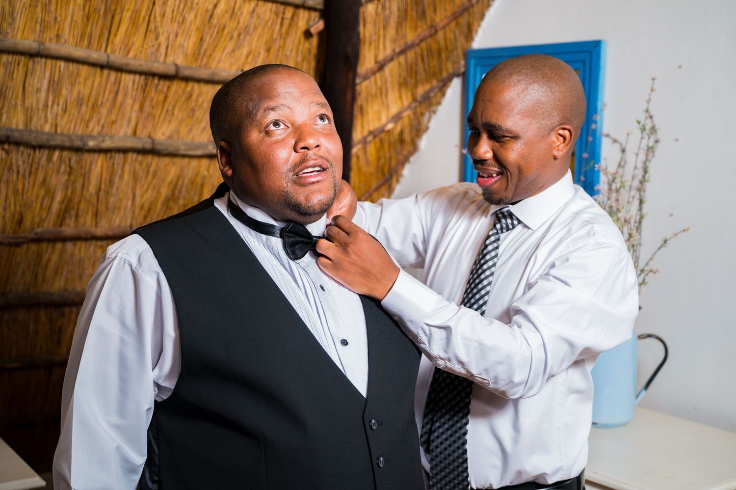 A man adjusts a bow tie on a groom wearing a black vest and white shirt at a wedding preparation.