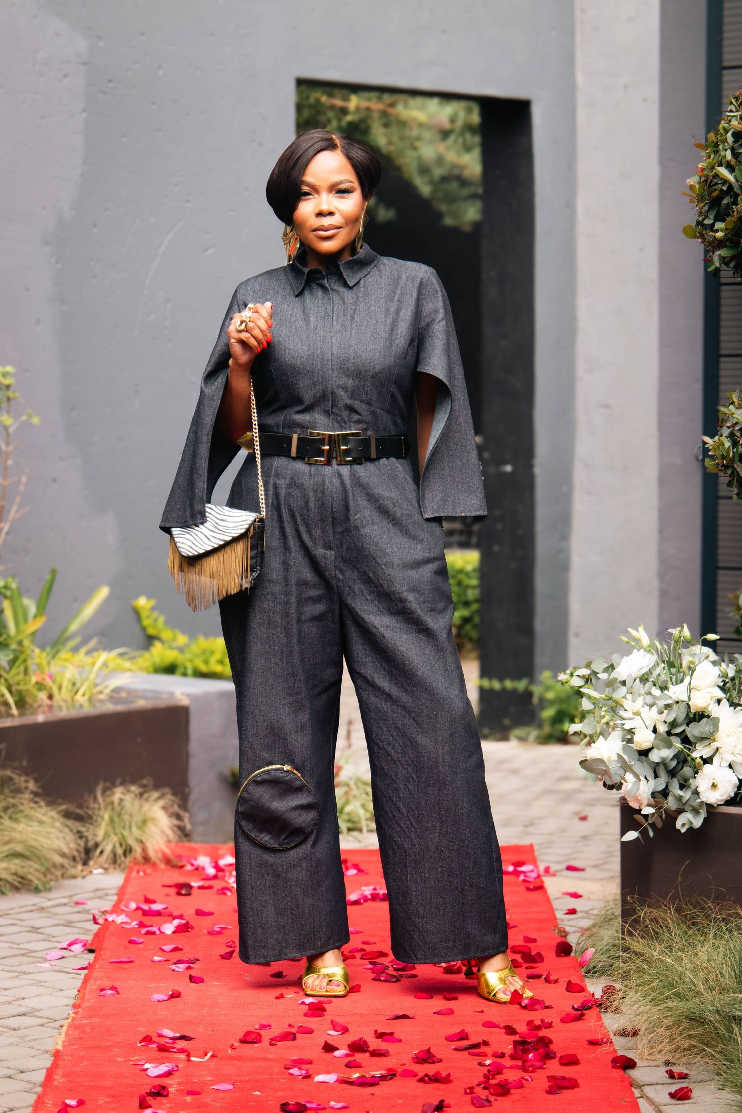 Woman in dark denim cape jumpsuit with wide-leg pants, black belt, and gold sandals posing on red carpet.