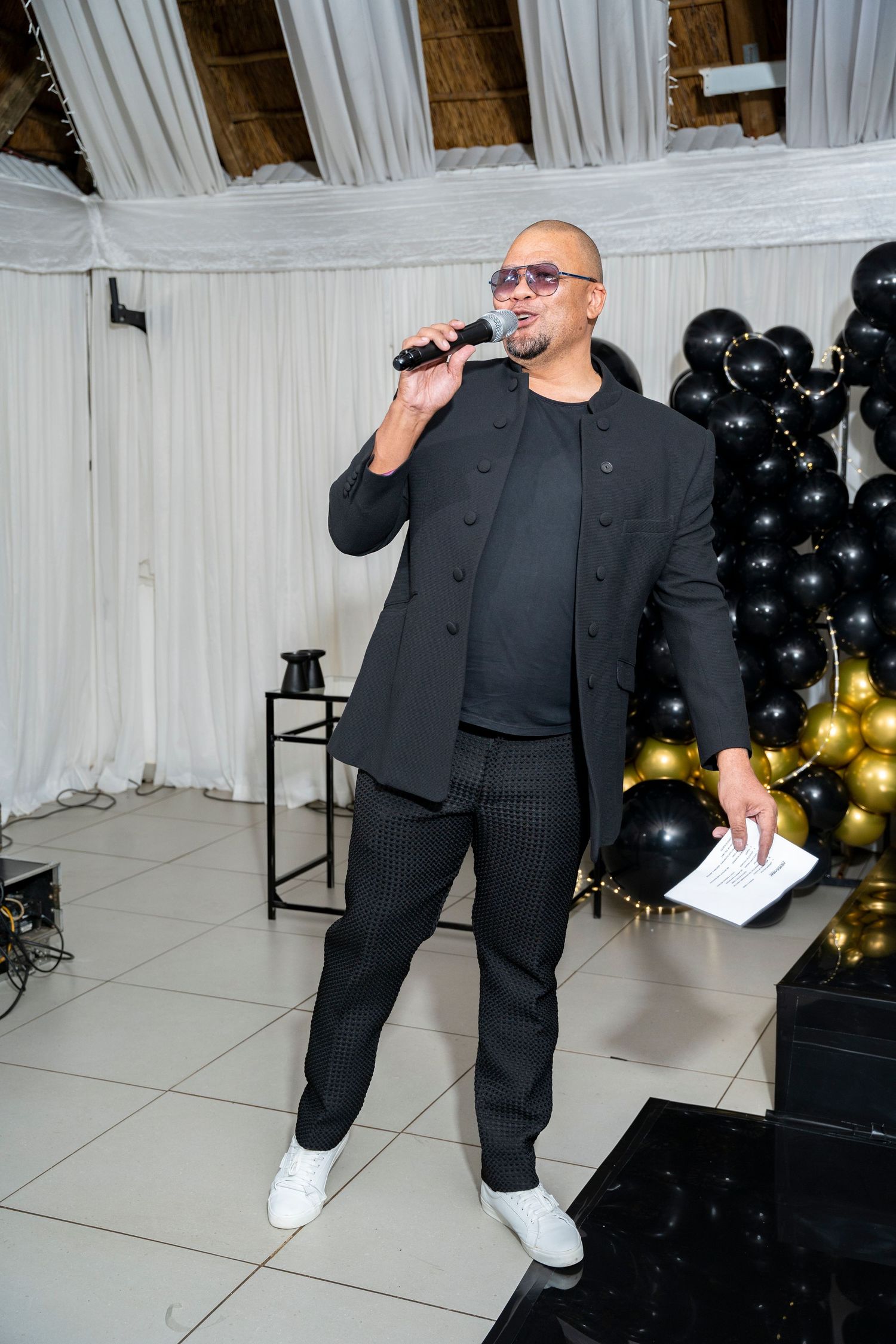 Stylish MC in black suit and sunglasses speaks into microphone at event with black and gold balloons.