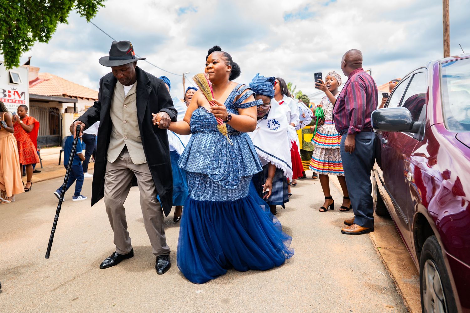 A woman in a blue gown walks with an elder man using a cane during a traditional celebration.