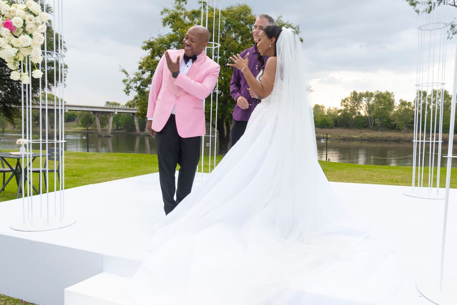 Groom in pink suit holds bride's hands at outdoor wedding altar near waterfront.