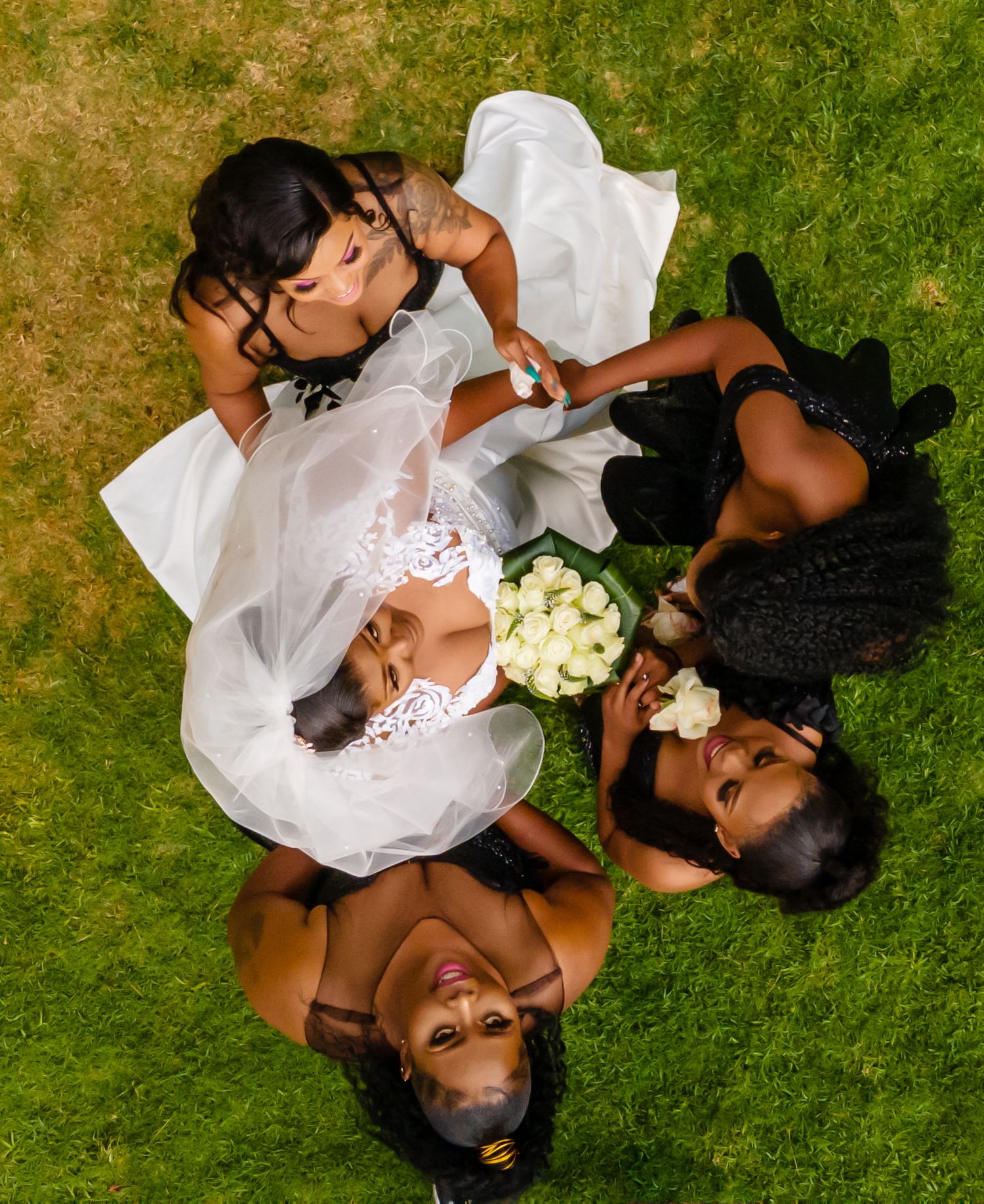 Aerial view of bride in white dress holding bouquet surrounded by bridesmaids lying on green grass.