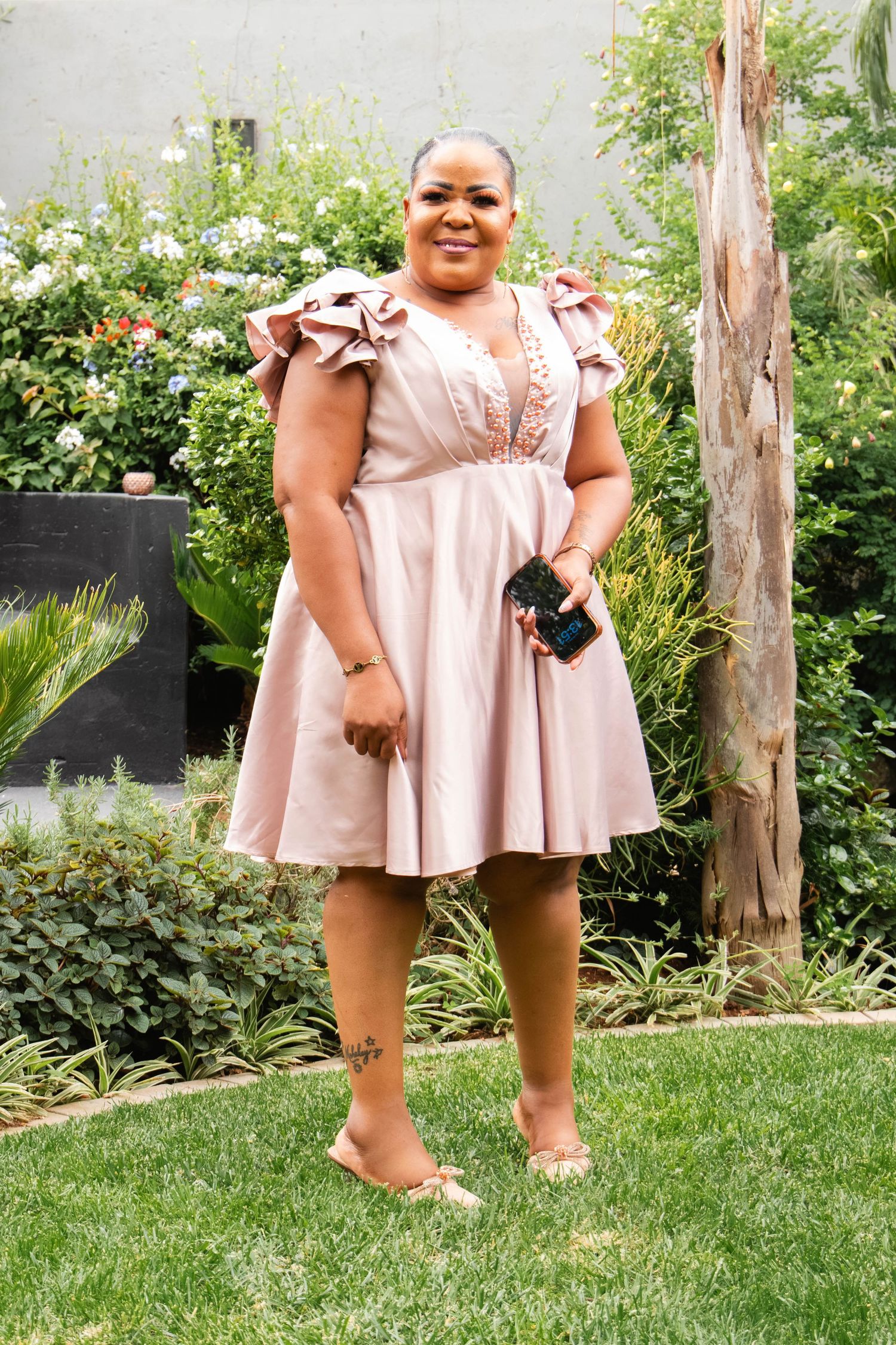Woman in pink ruffle dress holding phone, standing on green lawn with garden background.