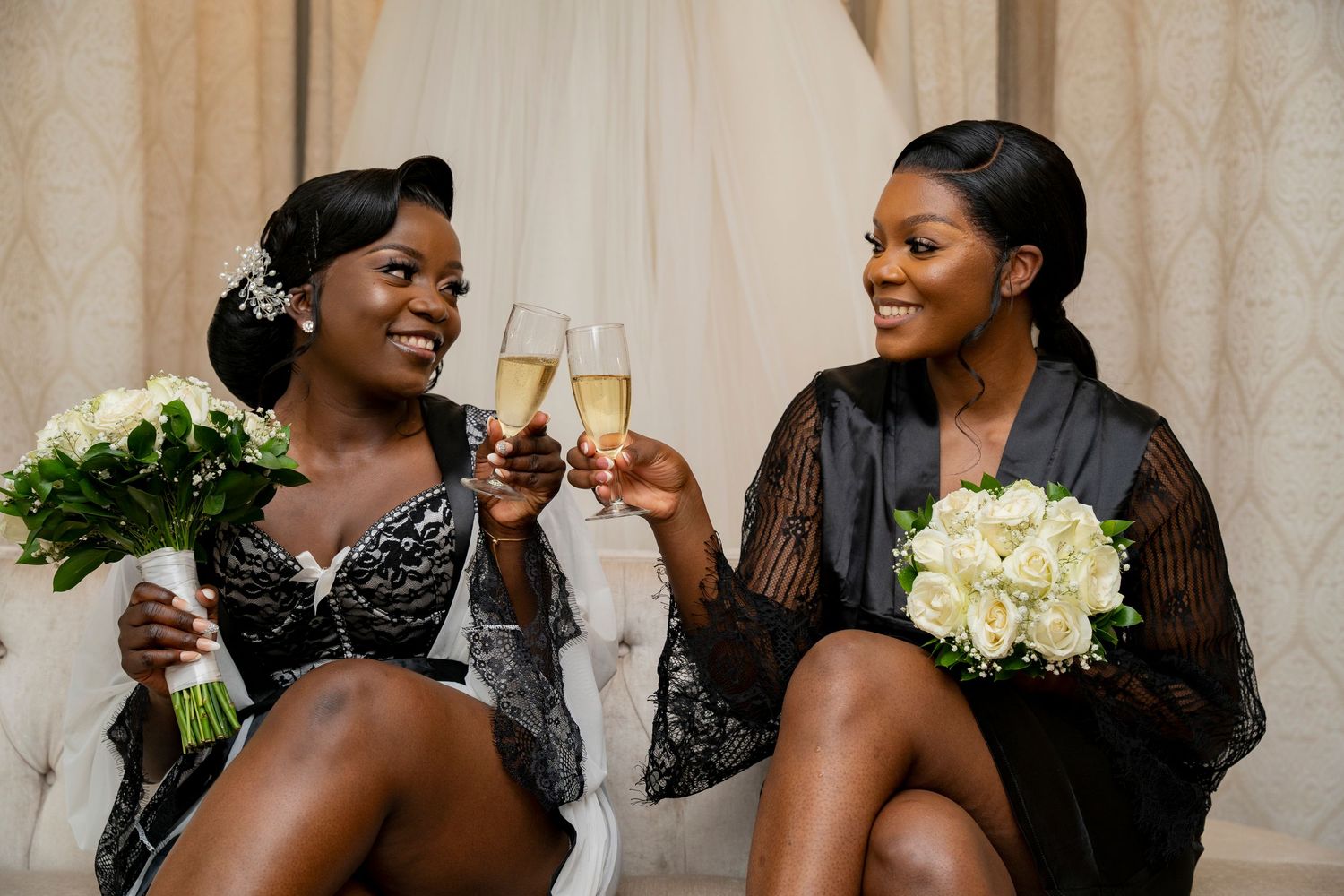 Two bridesmaids in black robes toasting champagne and holding white bouquets at a wedding celebration.