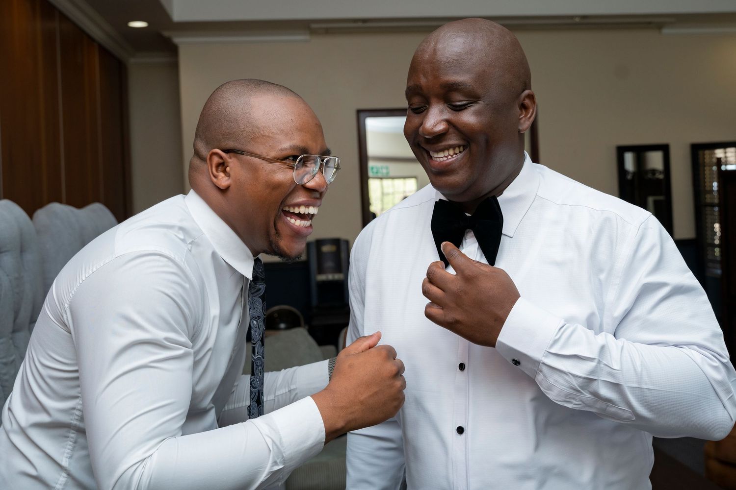 Two men in white dress shirts laughing and adjusting bow ties at a wedding celebration.