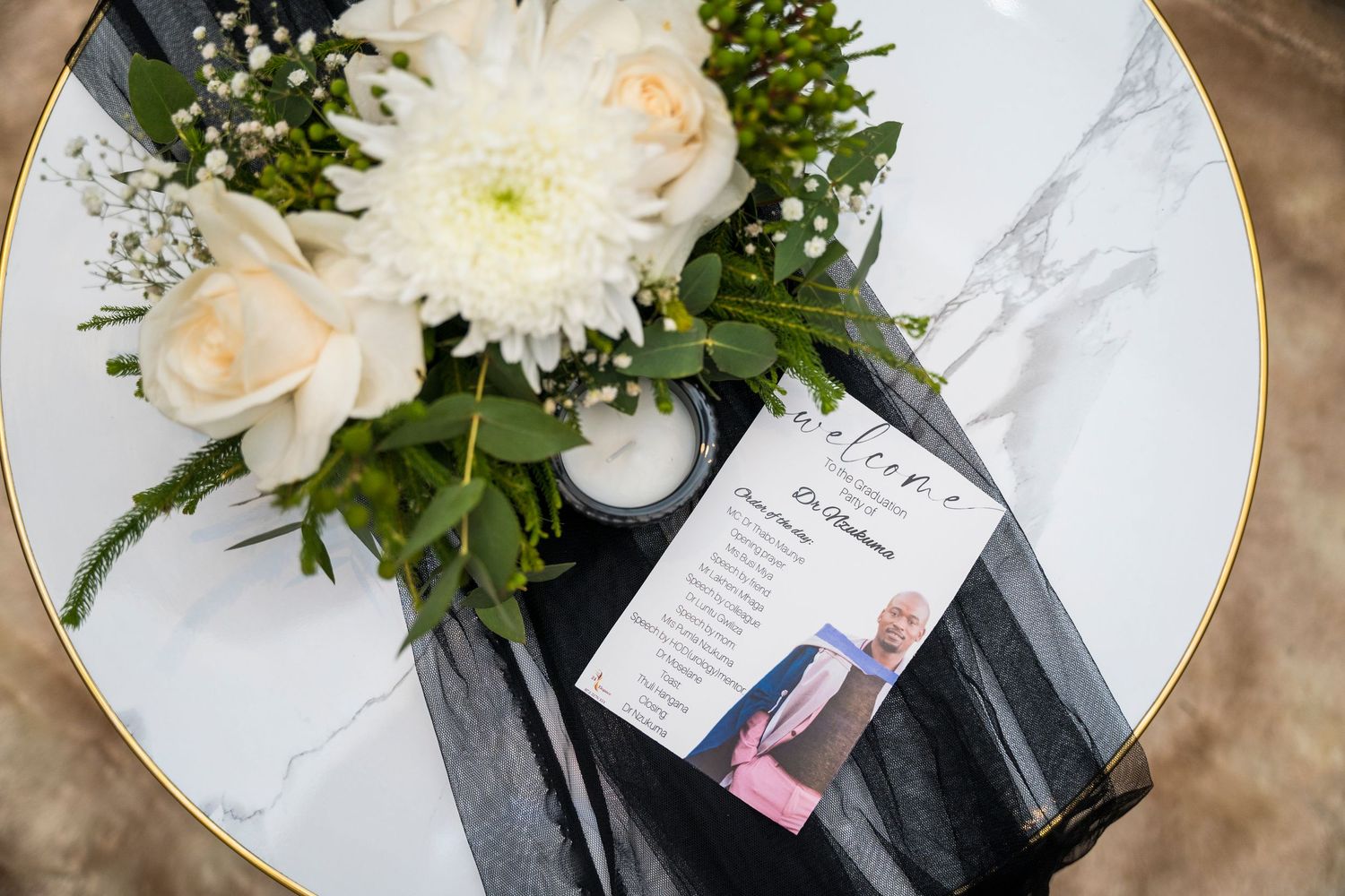 Elegant white floral centerpiece with roses and chrysanthemums beside a welcome card on a marble table.