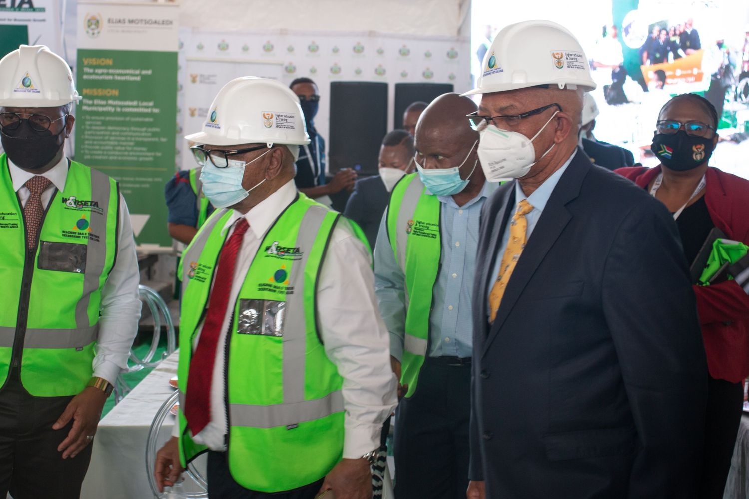 Officials in green safety vests and hard hats tour a South African infrastructure project site.