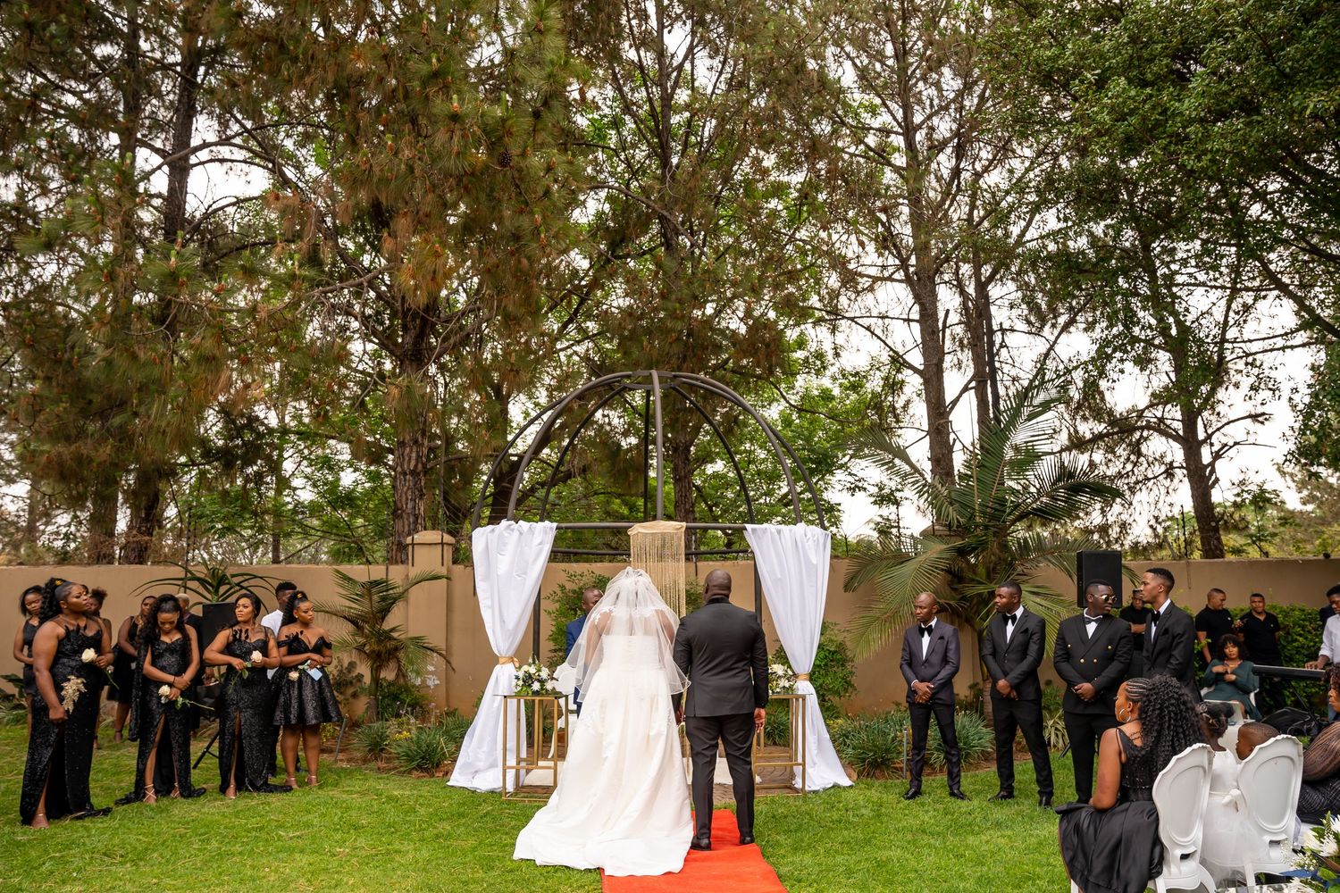 Wedding ceremony on green lawn with bride and groom at floral arch, guests seated on both sides.
