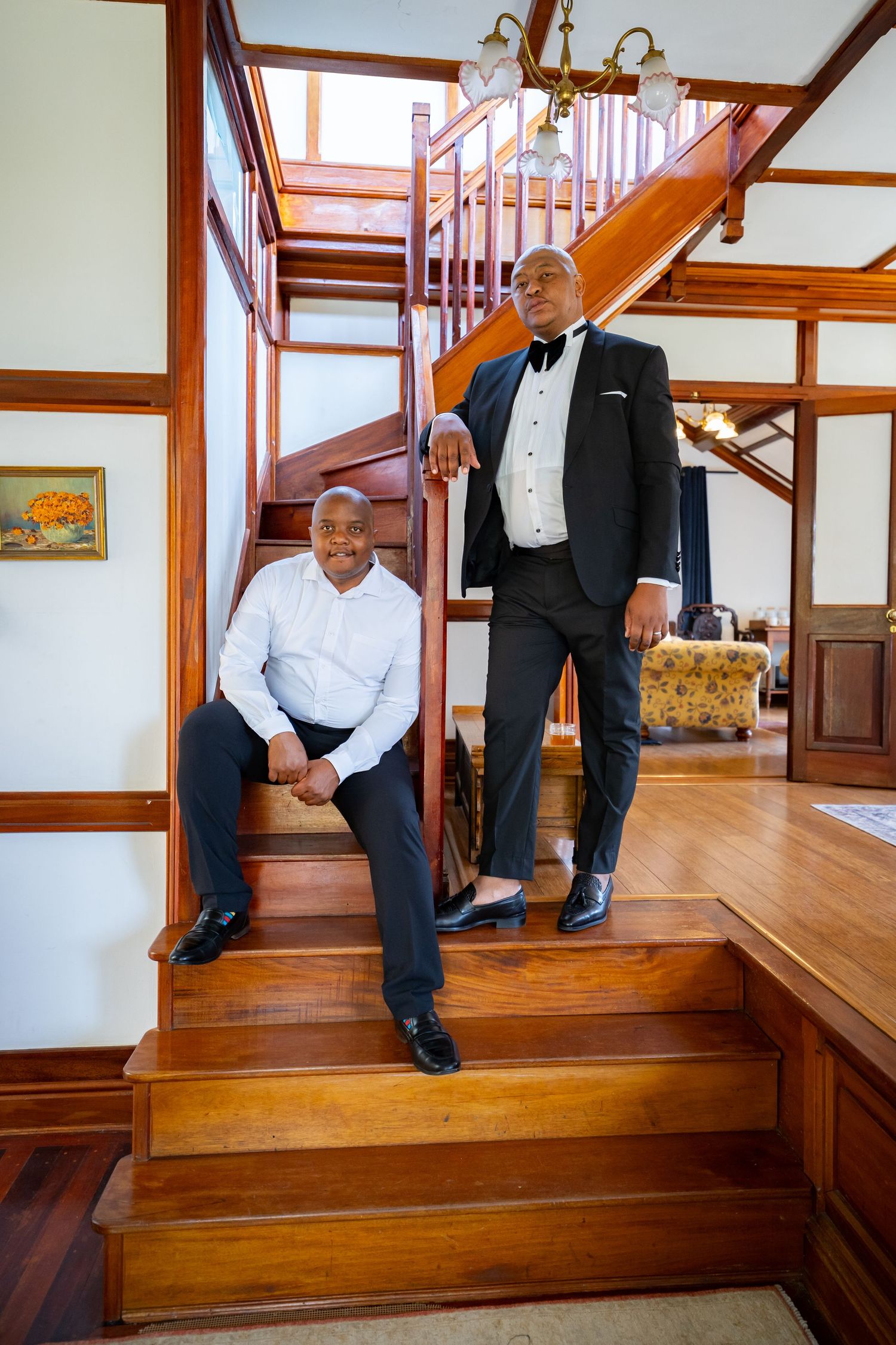 Two men in formal attire posing on wooden staircase, one seated and one standing in a tuxedo.
