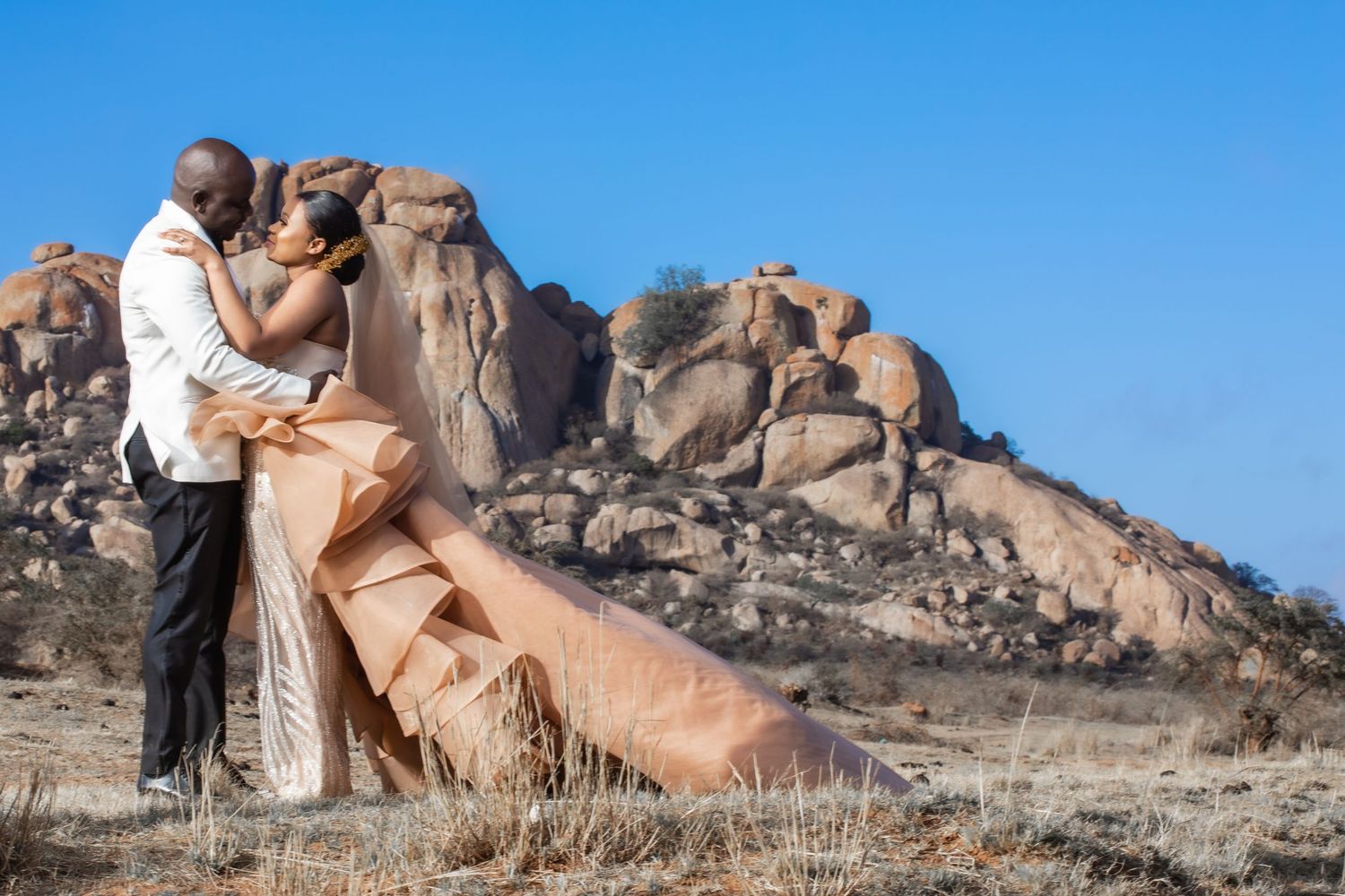 African couple embracing in wedding attire with dramatic rocky landscape under blue sky.