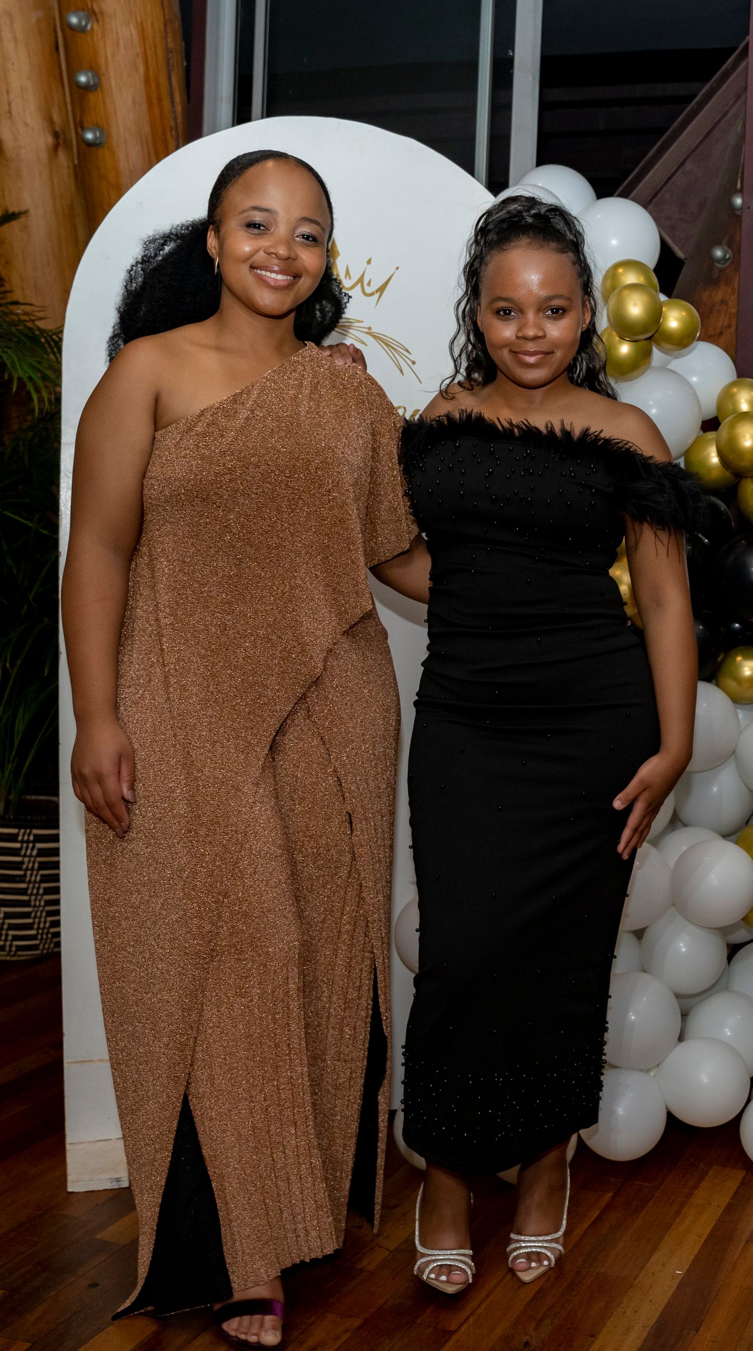 Two women in elegant evening gowns pose together at a formal event with gold and white balloon decorations.