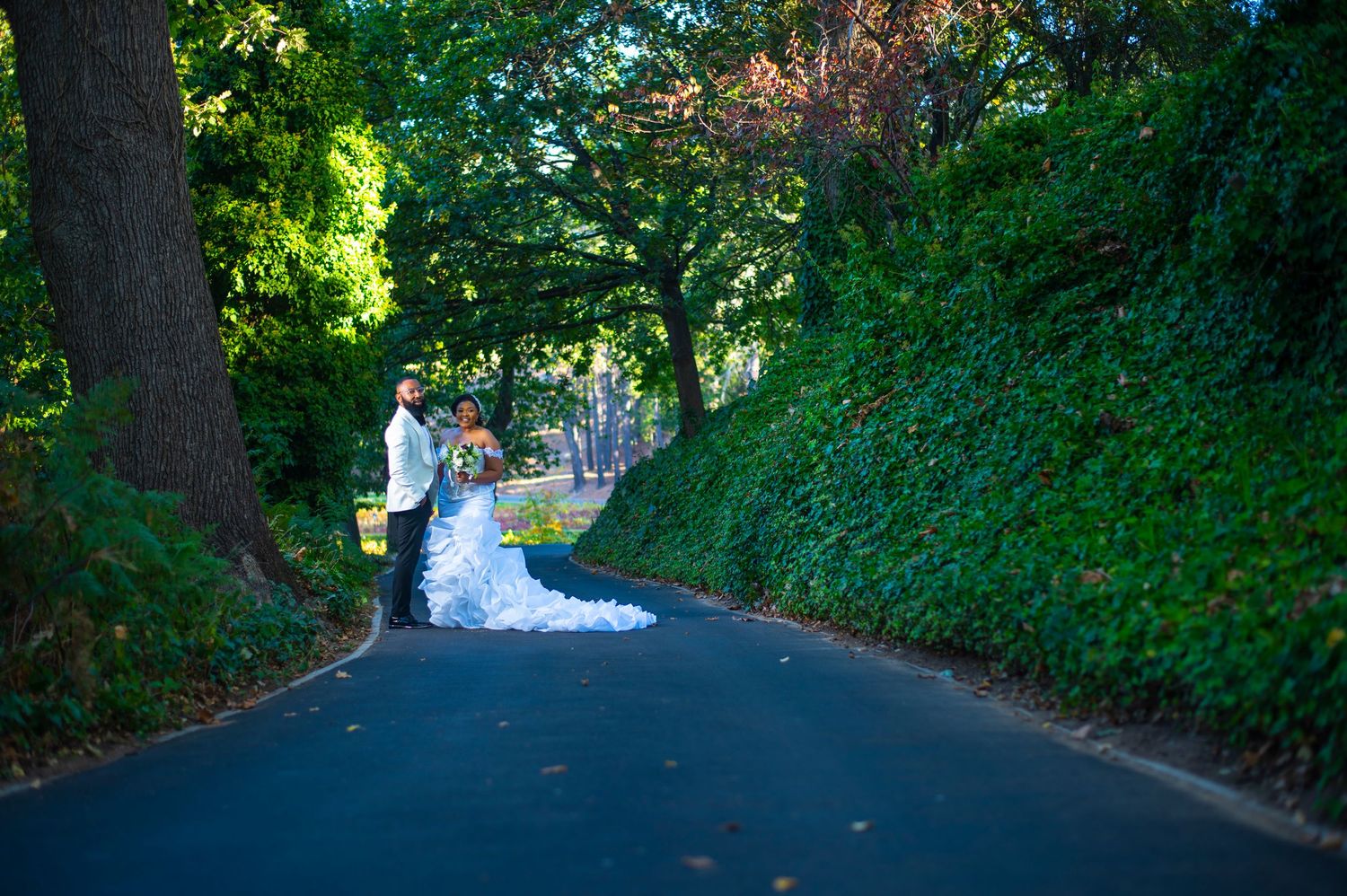 Bride and groom share romantic moment on tree-lined road with lush green surroundings.