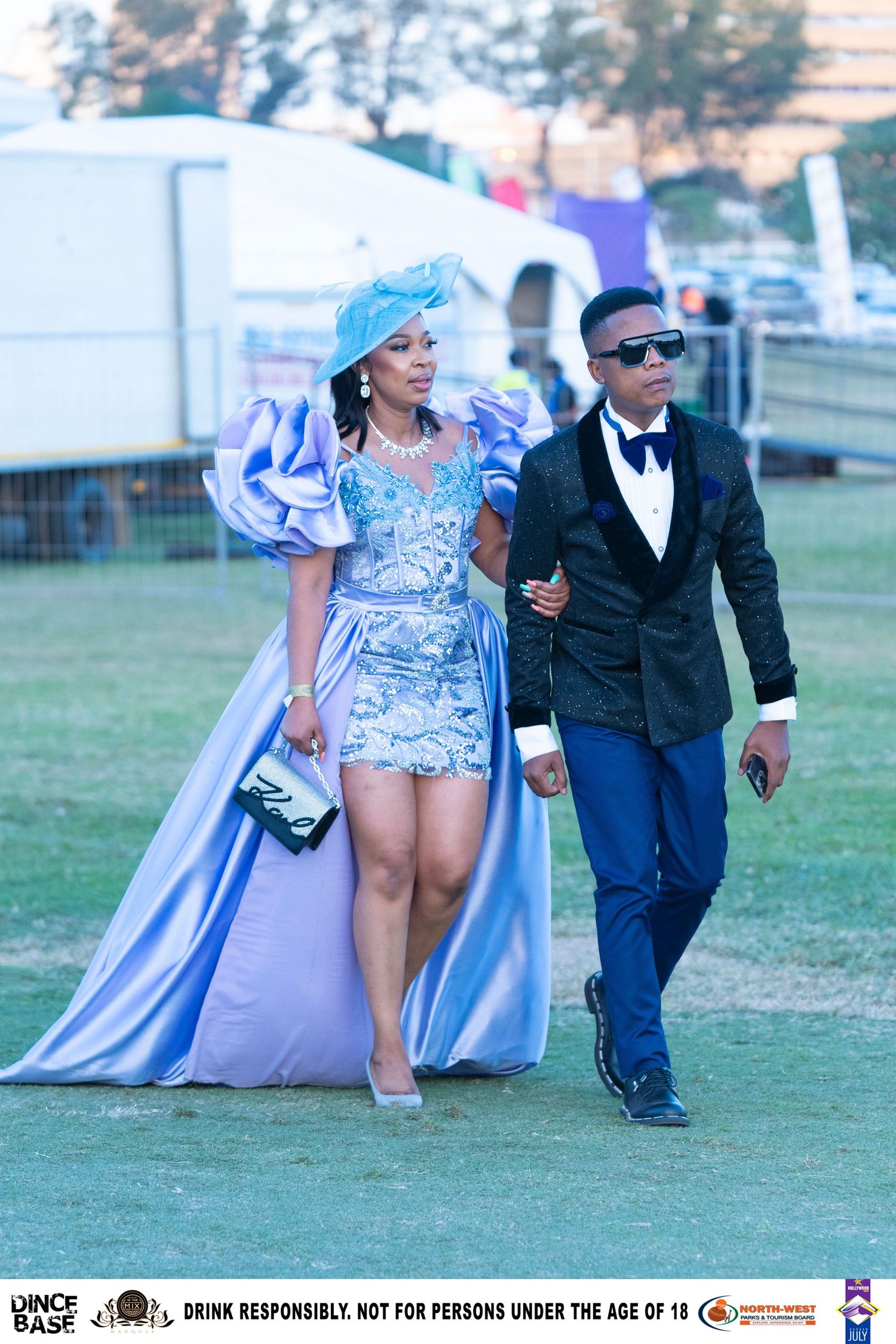 Stylish couple in coordinated blue formal attire walking at an outdoor racing fashion event.