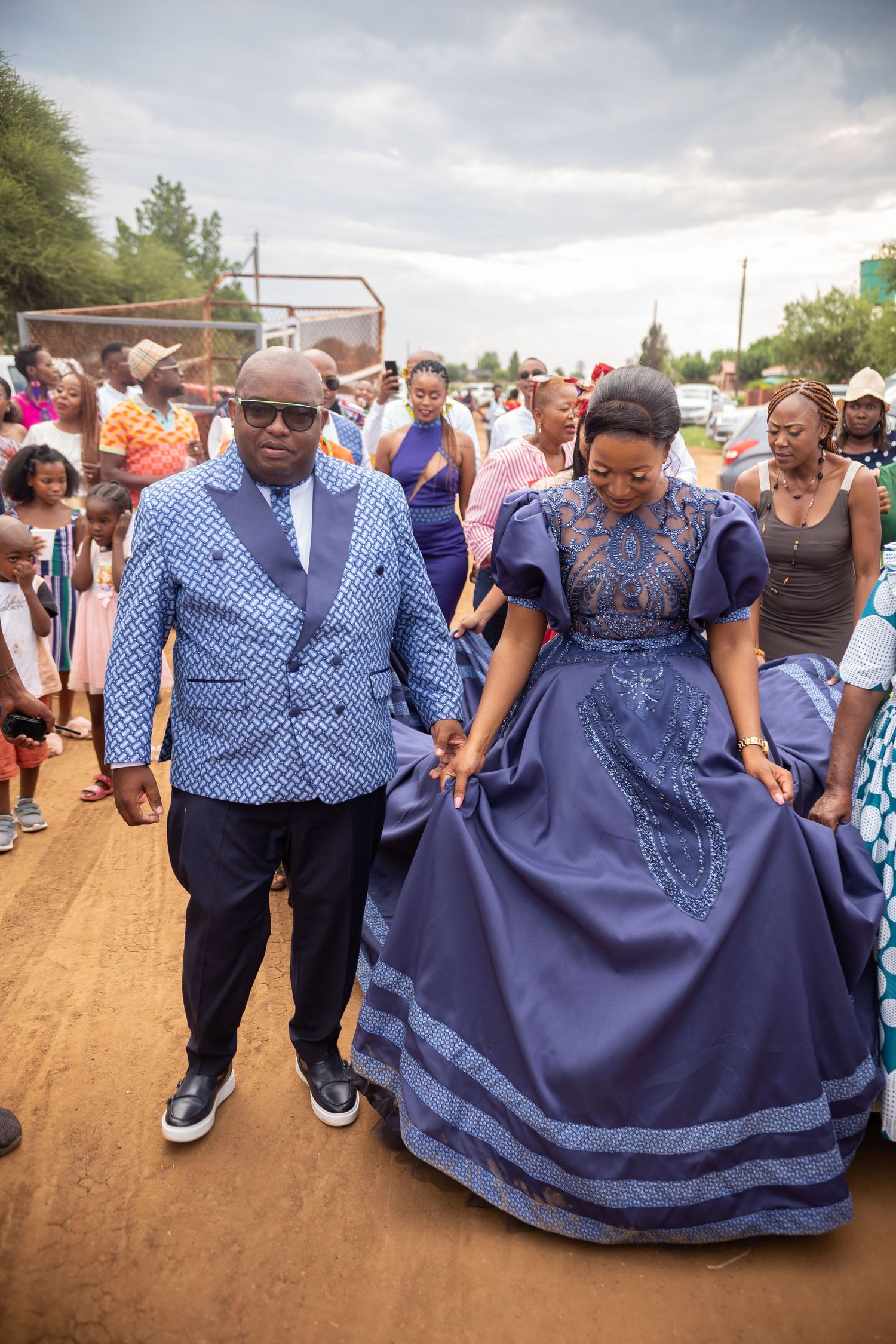 Couple in matching blue traditional African attire walking together at outdoor cultural wedding ceremony.