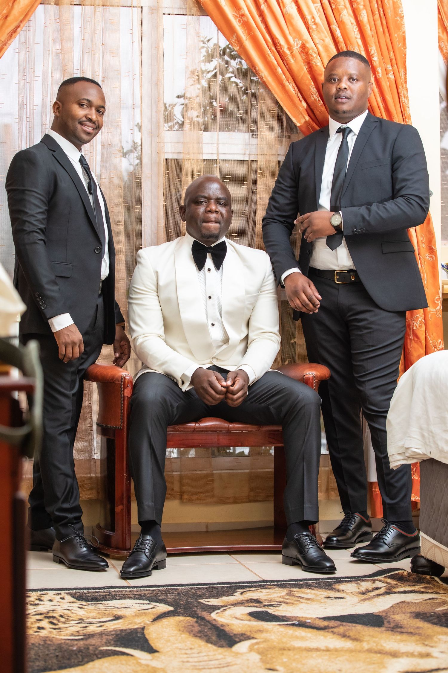 Groom in white tuxedo seated with two groomsmen in black suits posing indoors.