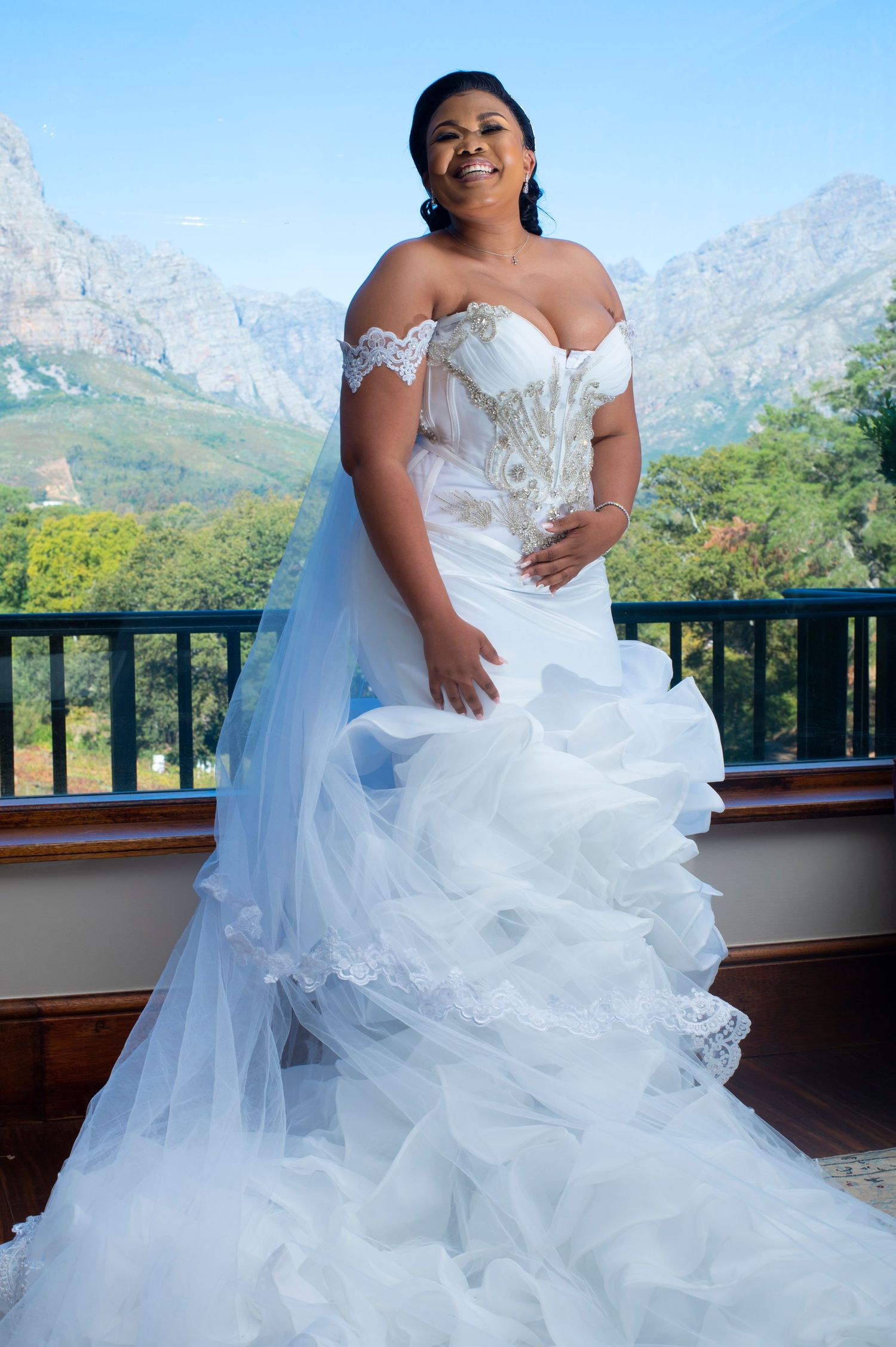 Radiant bride in off-shoulder mermaid wedding gown with lace details and veil, posing on balcony with mountain backdrop.