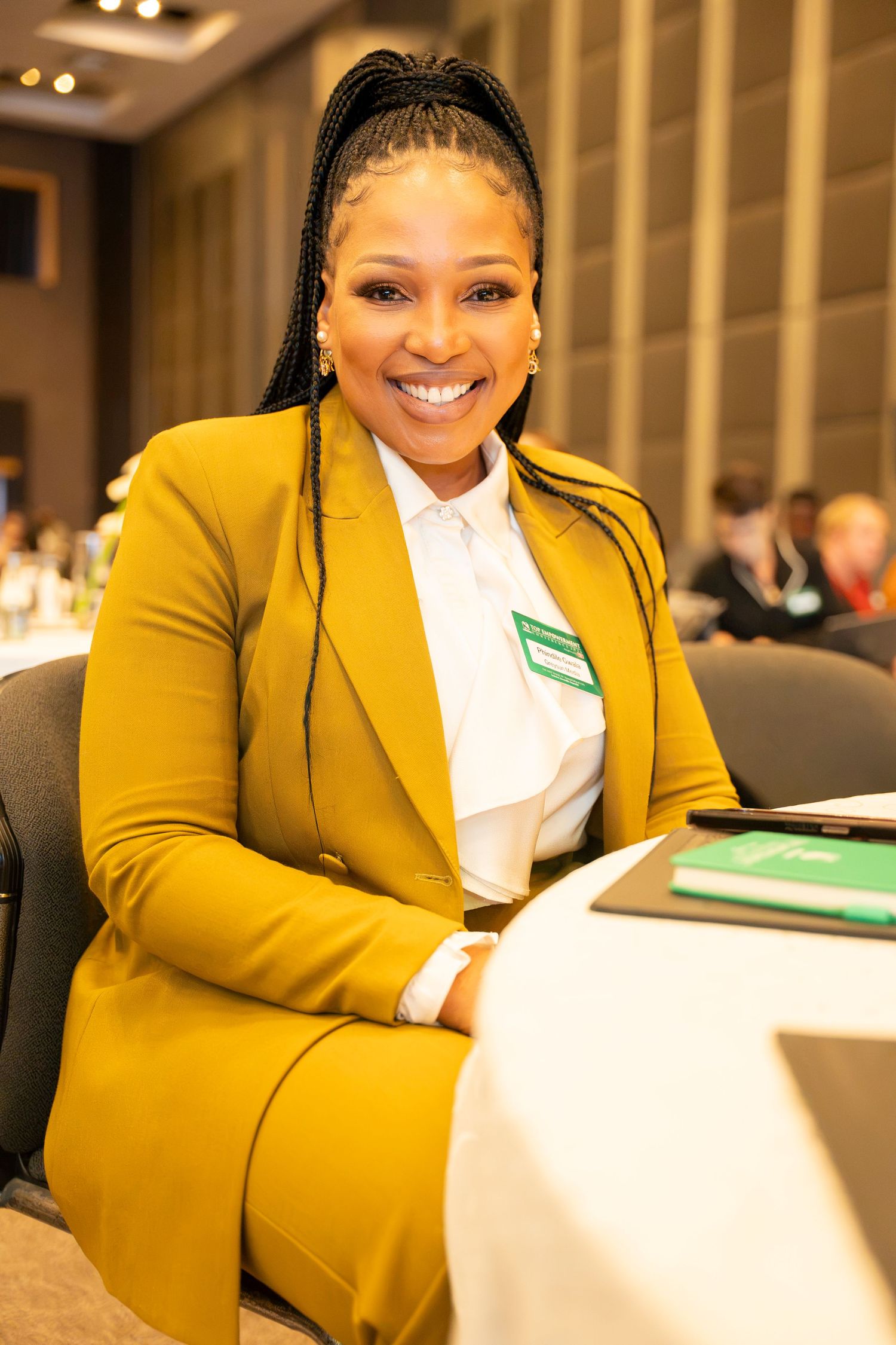 Smiling woman in yellow blazer with braids wearing a name badge seated at a conference table.