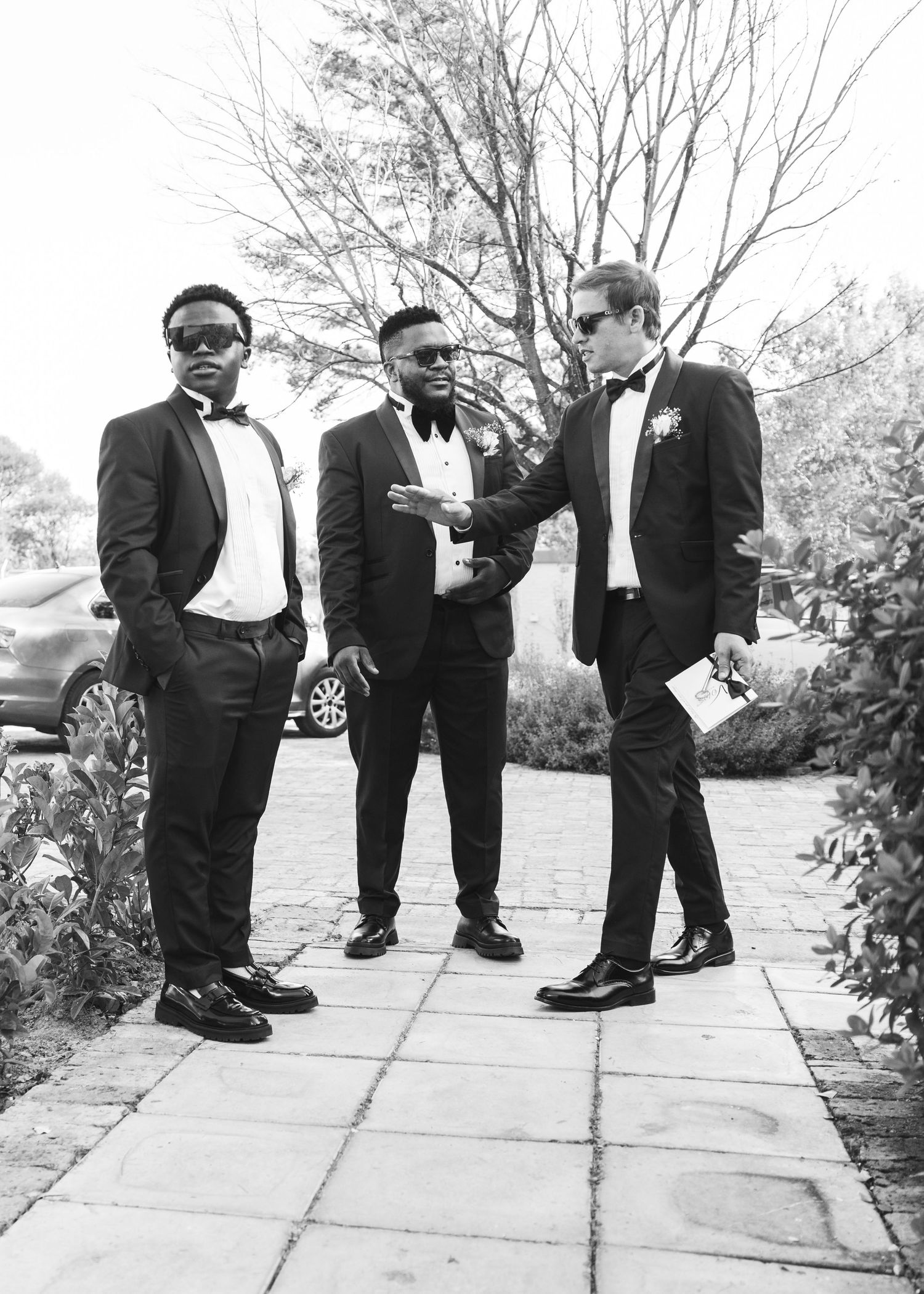 Three groomsmen in classic black tuxedos with bow ties conversing outdoors in stylish black and white photo.