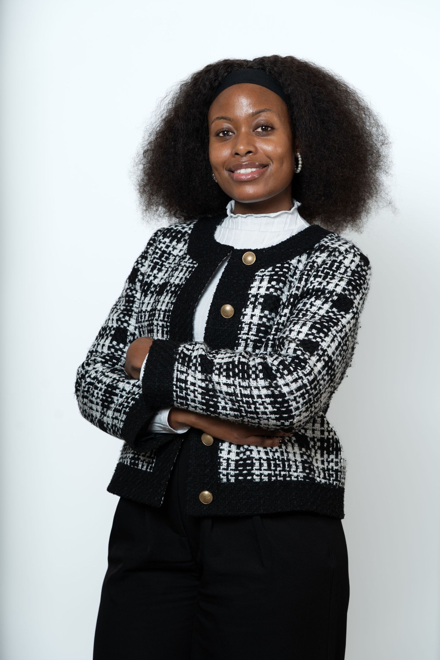 A smiling young Black woman with natural hair, wearing a plaid blazer, stands confidently with arms crossed against a white background.
