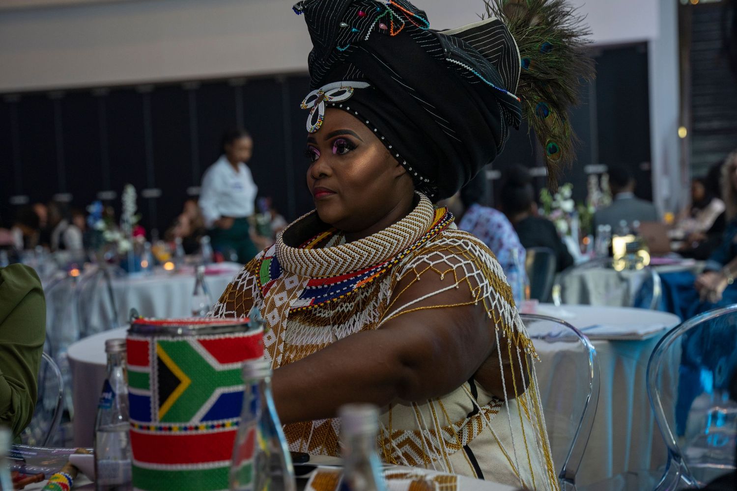 Woman in traditional Zulu beaded attire and headdress at a formal event with South African flag decor.
