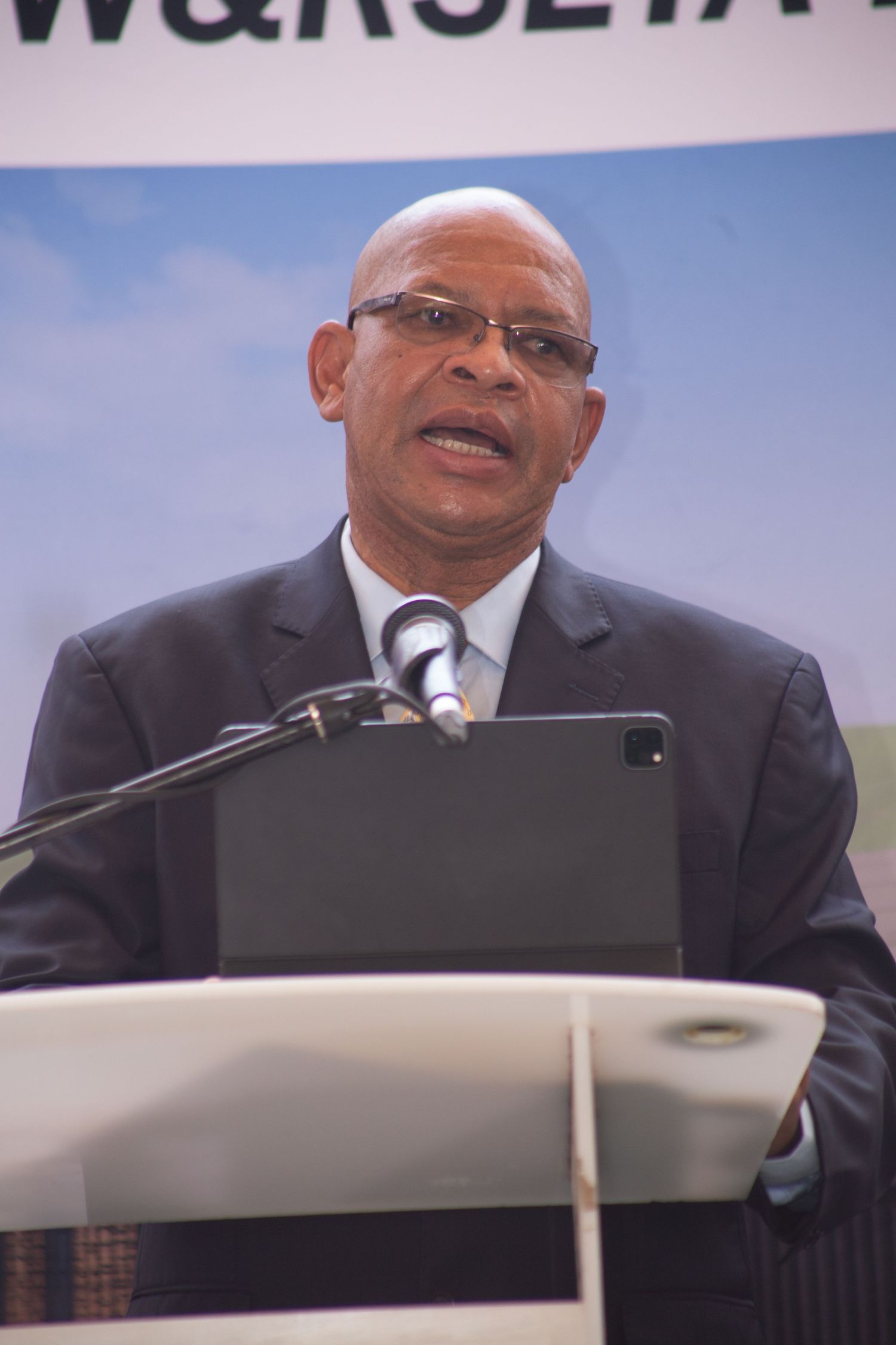 A man in a dark suit and glasses speaks at a podium with a microphone at a formal event.