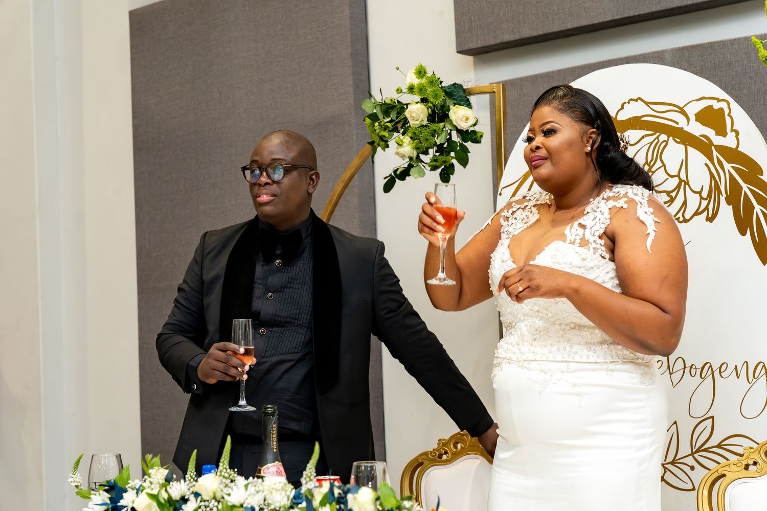 Newlyweds raise champagne glasses for toast at decorated wedding reception sweetheart table.