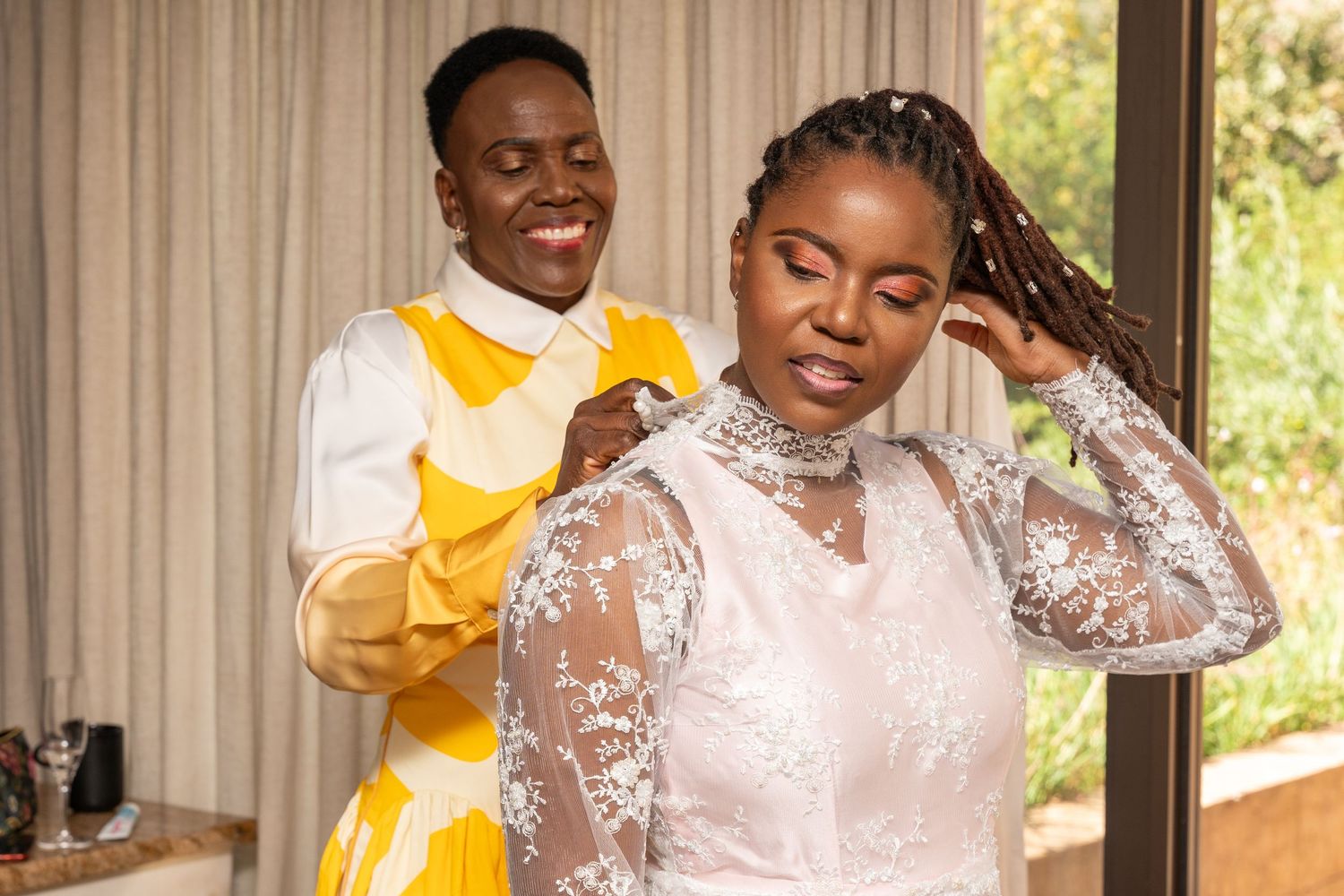 Bridesmaid in yellow dress helping bride in white lace gown with hair before wedding ceremony indoors.