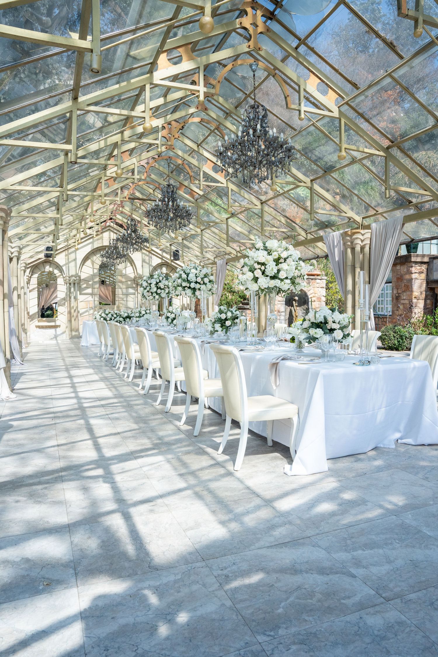 Elegant wedding reception venue with glass ceiling, white floral centerpieces, and chandelier decor.