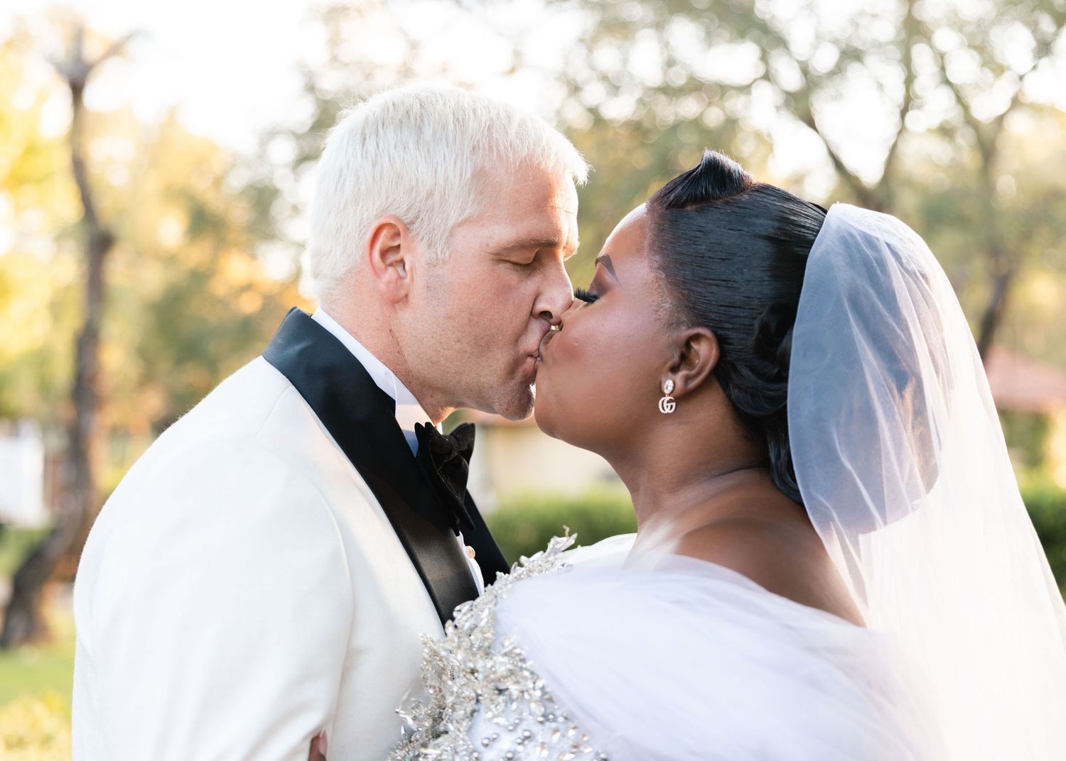 Interracial newlywed couple shares a romantic first kiss outdoors after their wedding ceremony.