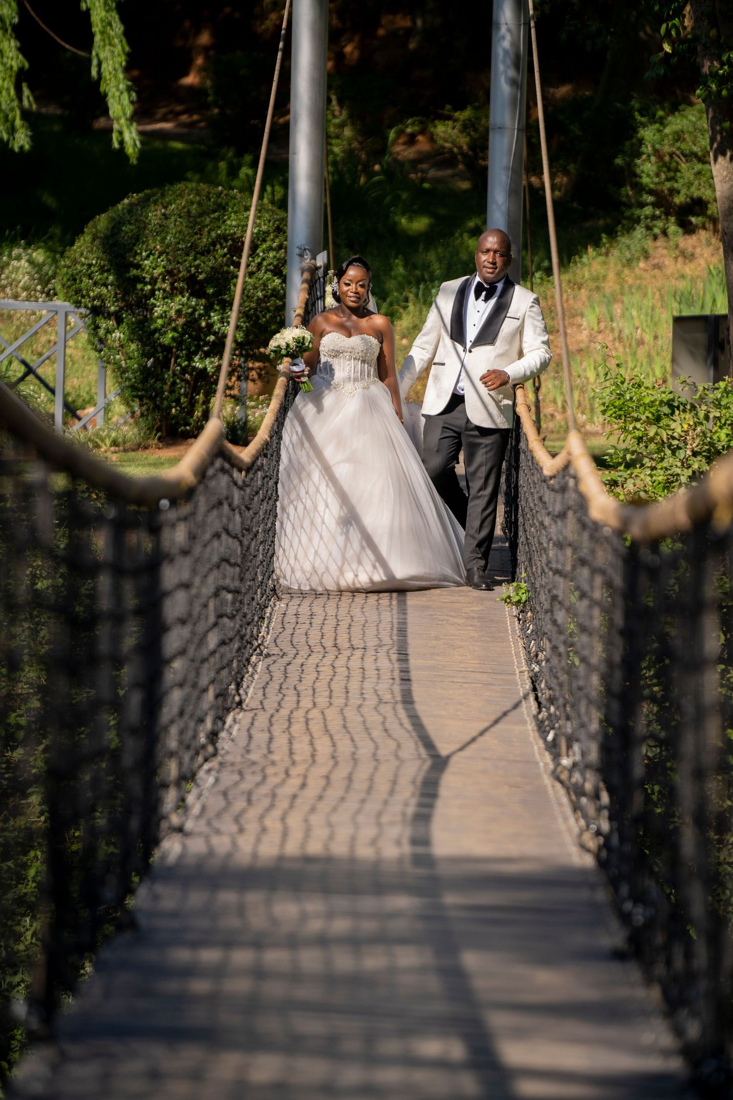 Bride and groom walking hand in hand across a wooden suspension bridge outdoors.