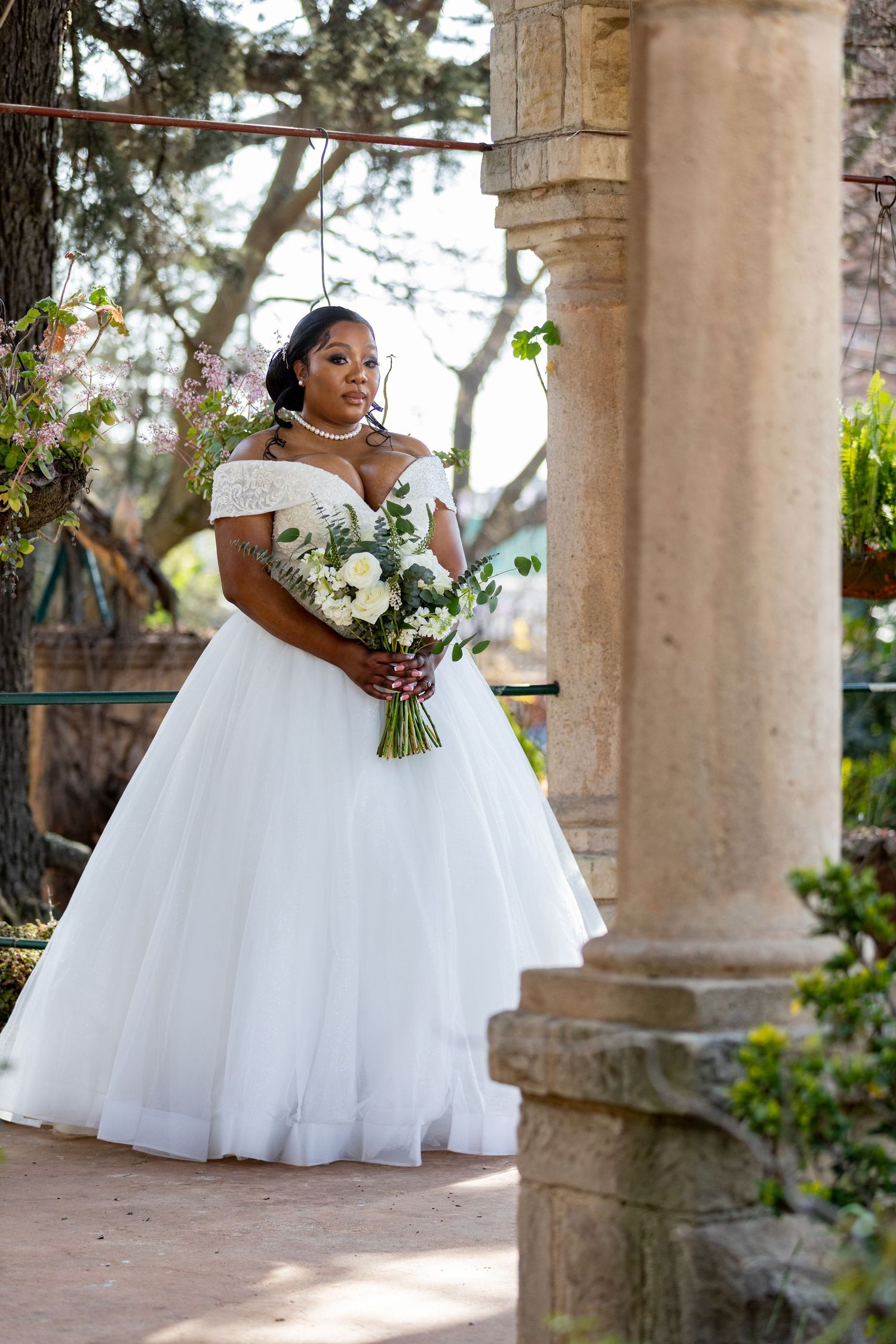 Elegant bride in white ball gown with bouquet standing between stone columns in garden setting.