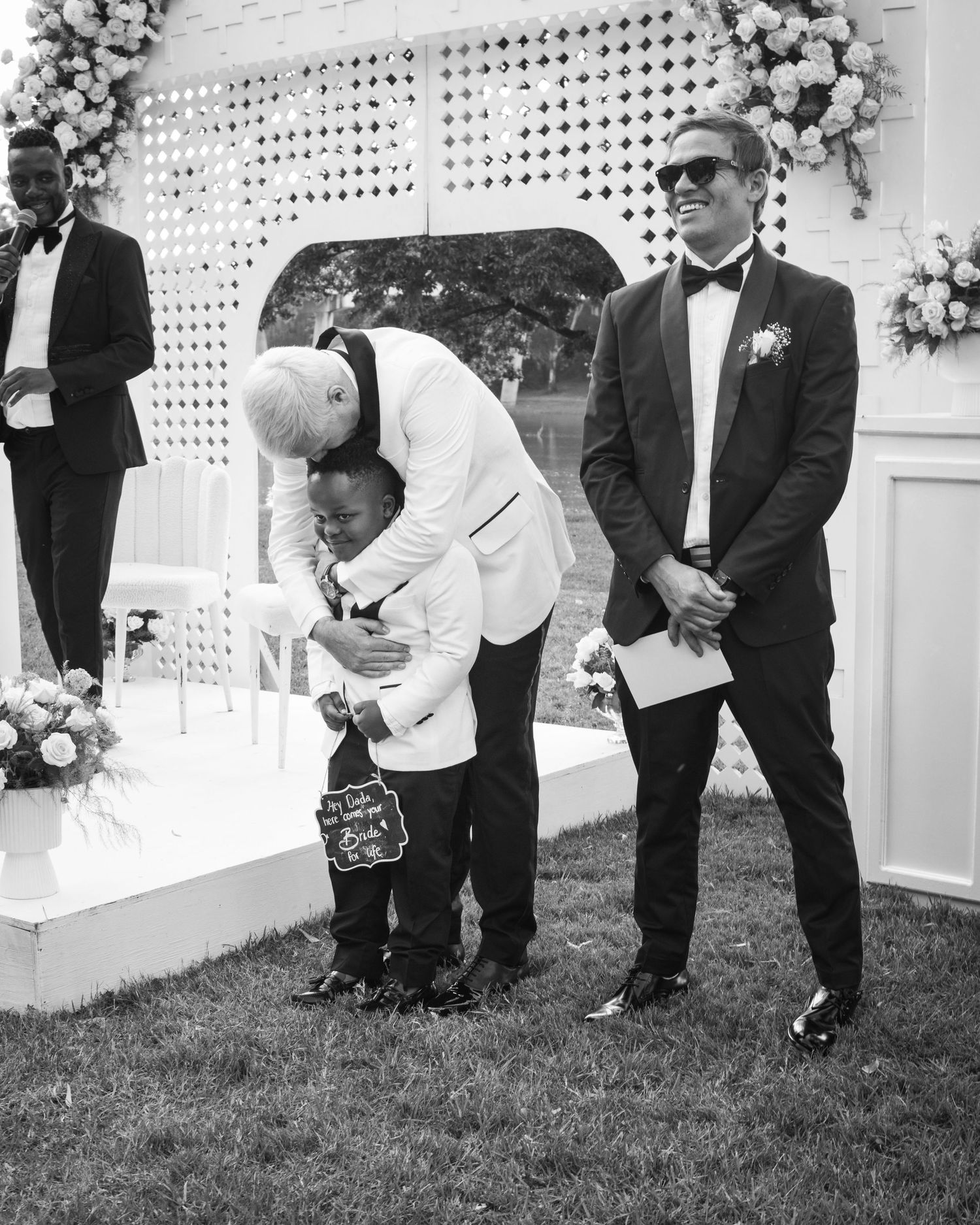 Black and white photo of a groom in a white tuxedo bending down to greet a young ring bearer.