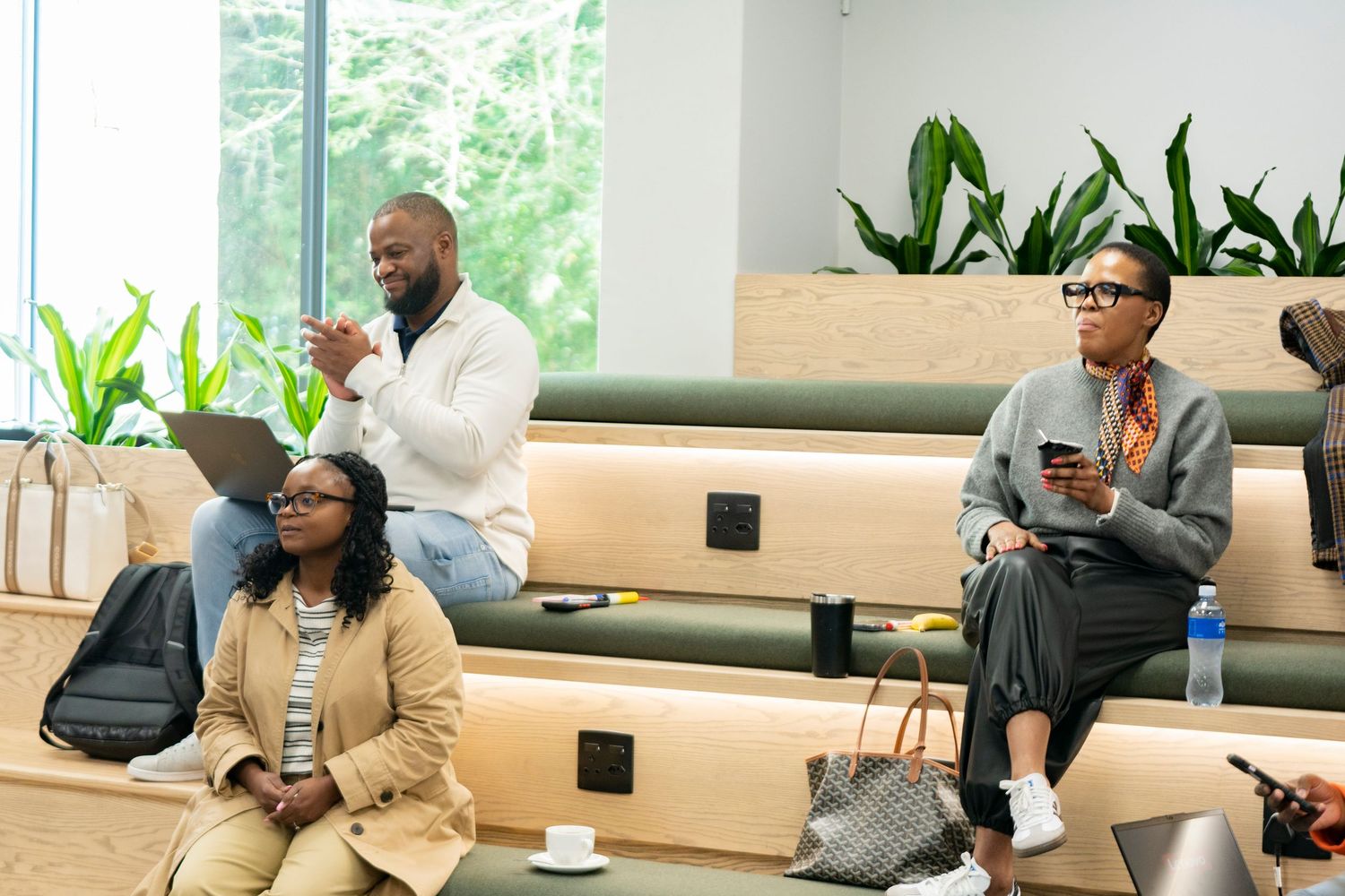 Three Black professionals sit in a modern conference room, two using laptops and one holding a smartphone.
