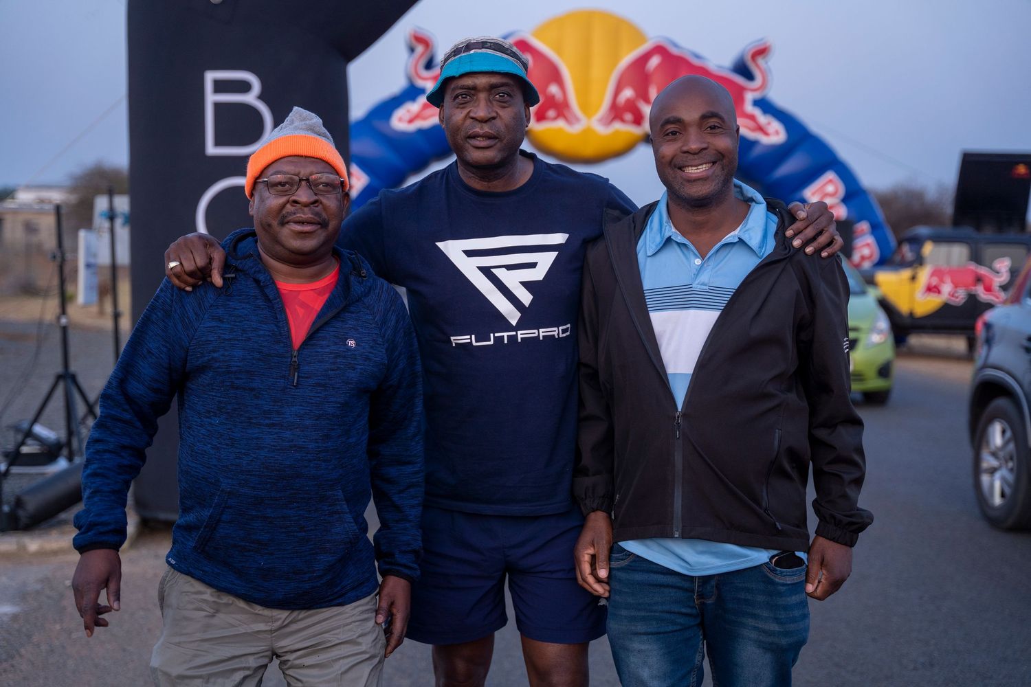 Three smiling men pose together at a Red Bull outdoor sporting event at dusk.