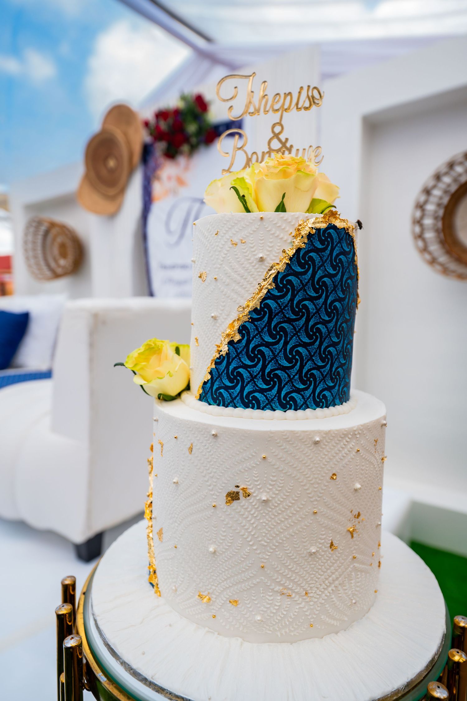 A two-tier white and blue African print wedding cake with gold accents and floral decorations.