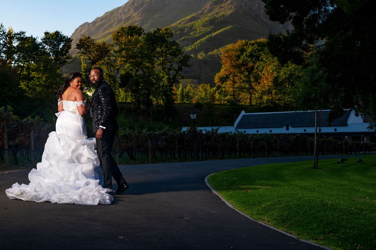 Bride and groom walking hand in hand along scenic vineyard path with golden mountain sunset backdrop.