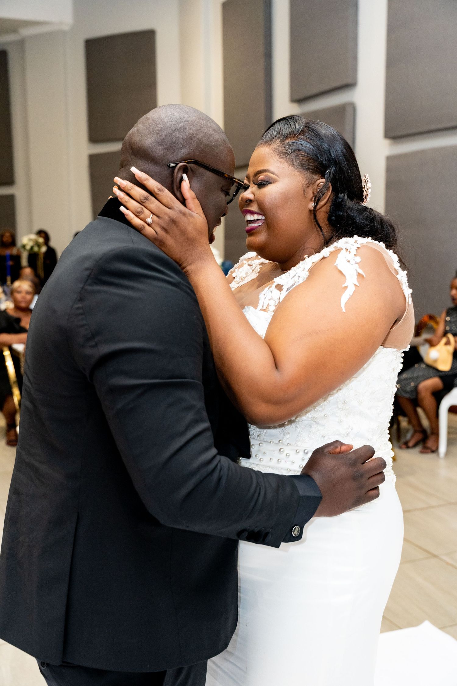 Bride in lace wedding gown laughs joyfully with groom during first dance at wedding reception.
