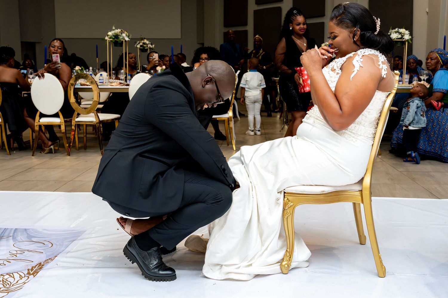 Groom kneeling before bride in white gown at elegant wedding reception, sharing an emotional moment.