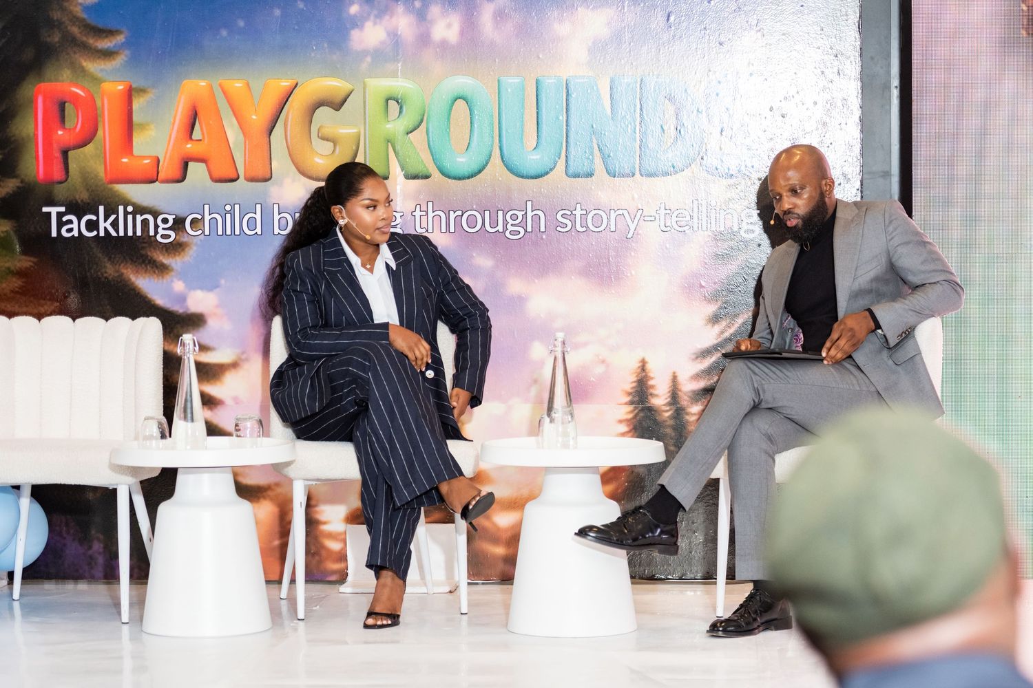 Two speakers at a Playground child literacy event discuss storytelling on a bright white stage.