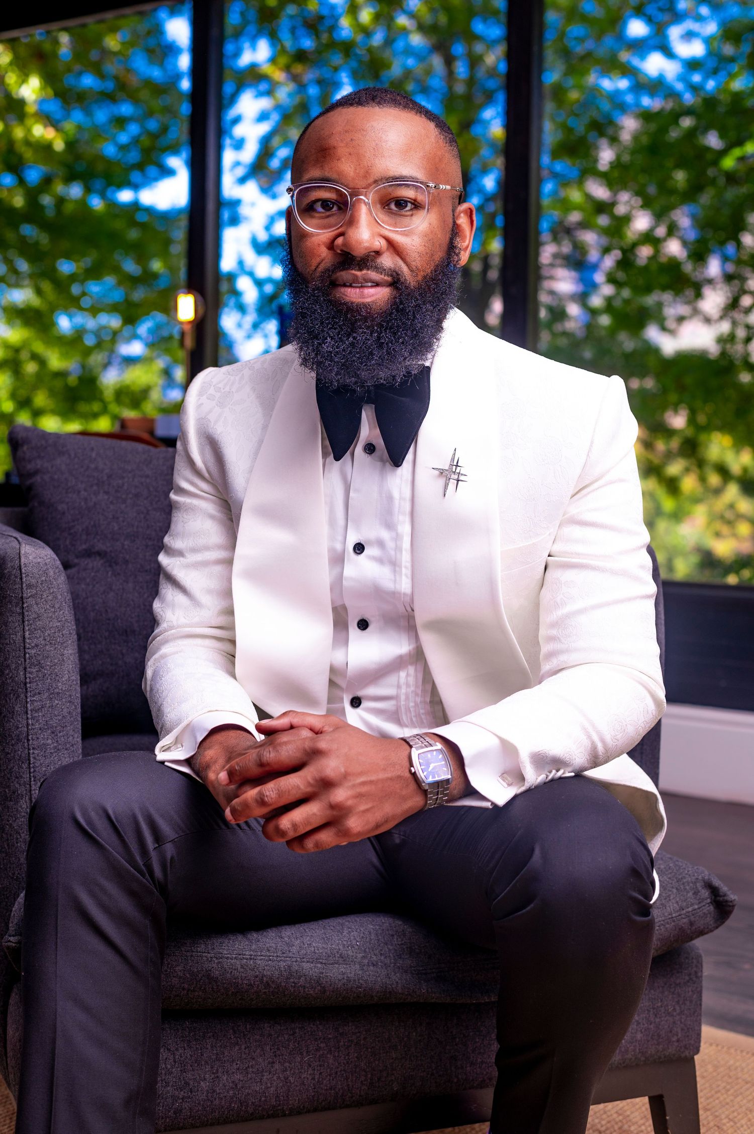 Stylish bearded groom in white tuxedo jacket with bow tie seated by green trees.