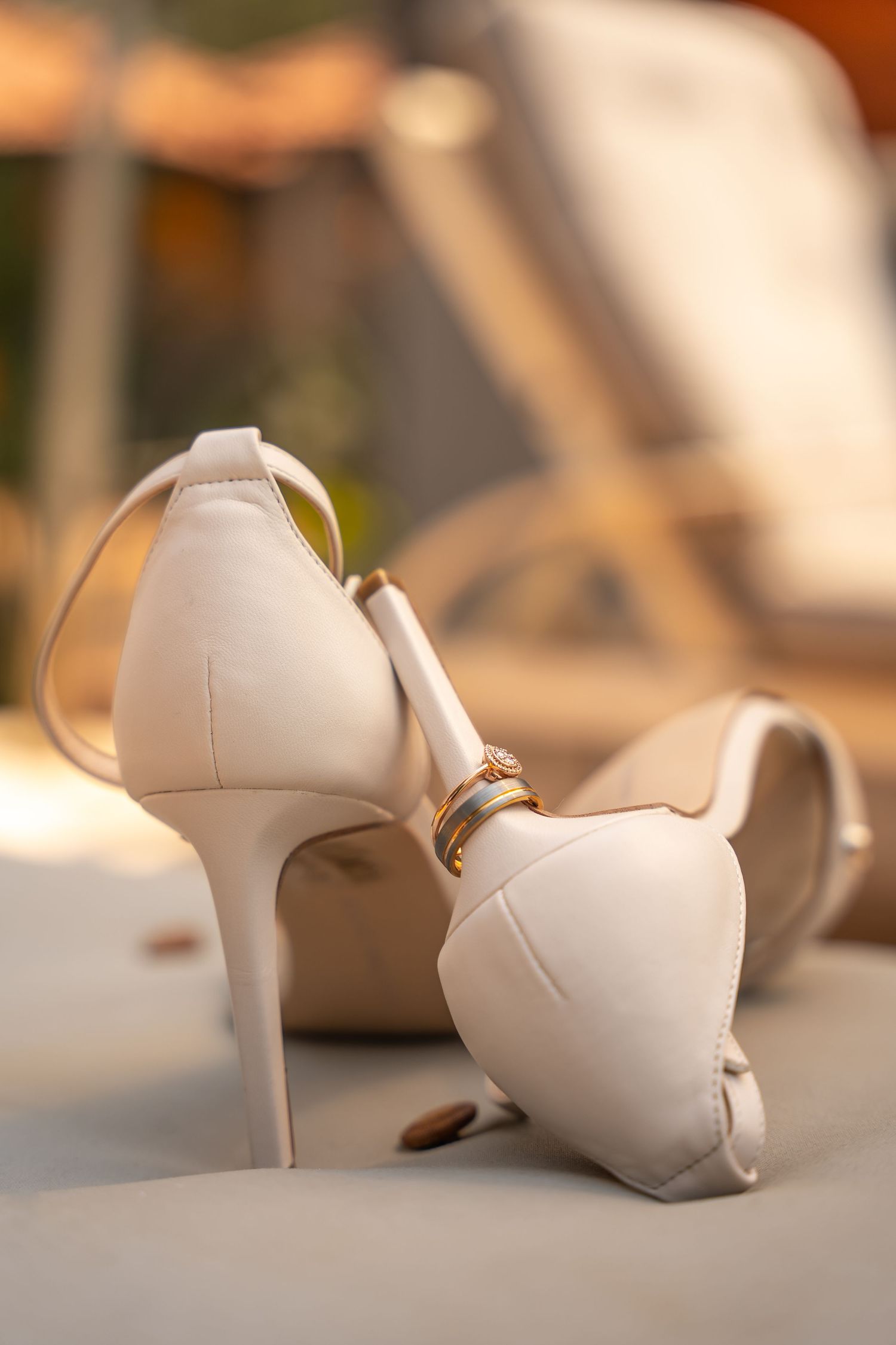 Elegant cream bridal heels with gold wedding rings resting on the back of the shoes outdoors.