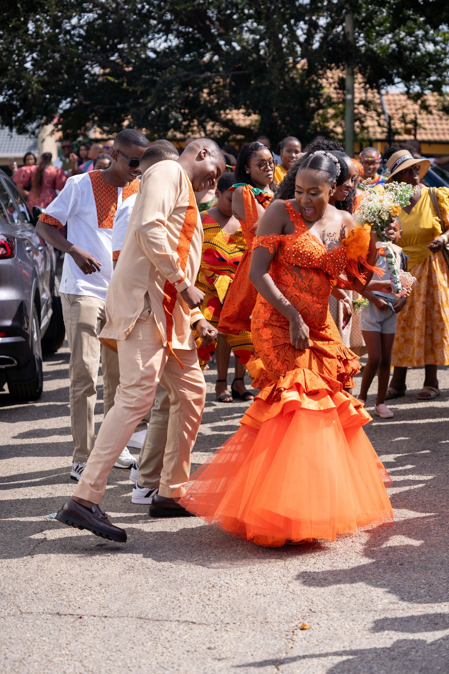 Bride in vibrant orange African traditional wedding dress dancing with groom on street celebration.