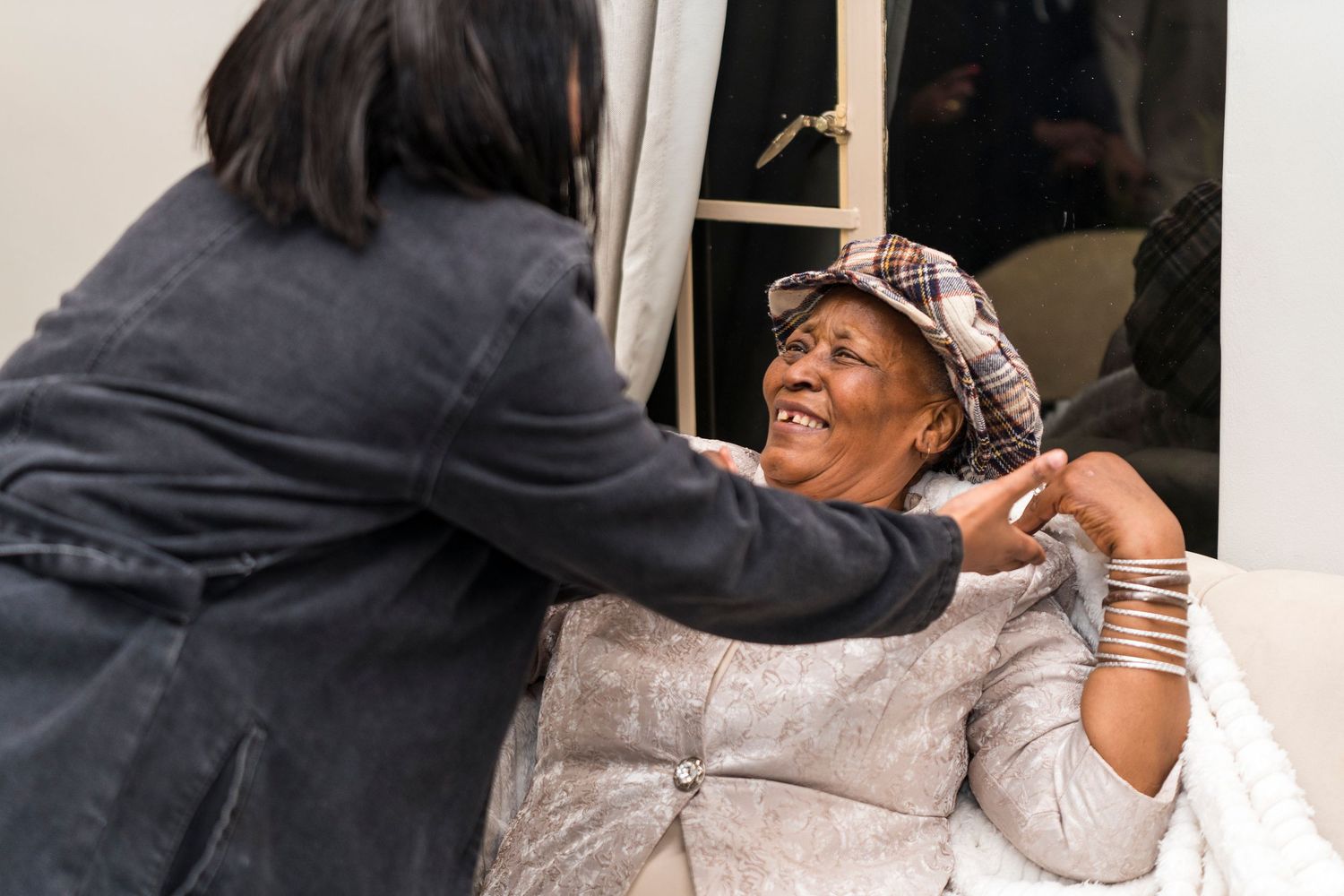 Woman in patterned headwrap smiles warmly as another person adjusts her outfit at an indoor event.