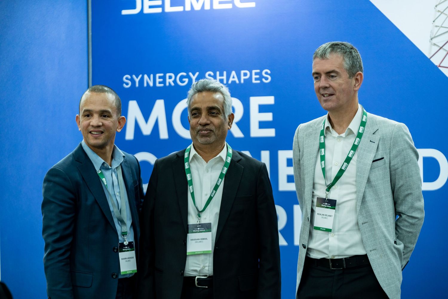 Three men smile for a photo in front of a Jelmec synergy shapes the world branded backdrop.