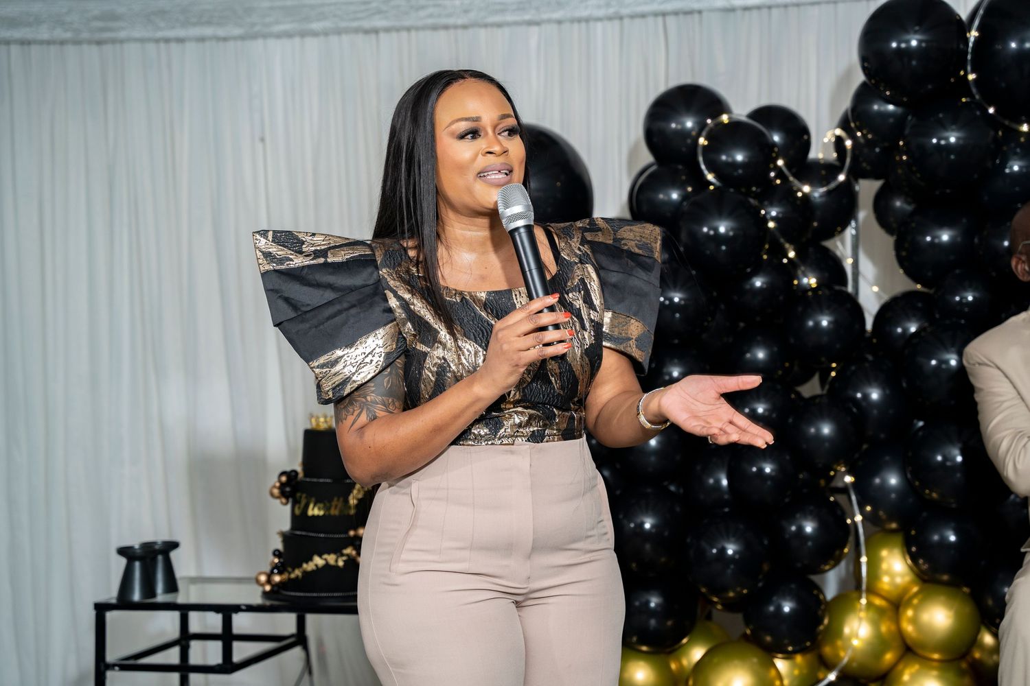 Woman with dramatic shoulder detail speaks into microphone at elegant event with black balloon backdrop.