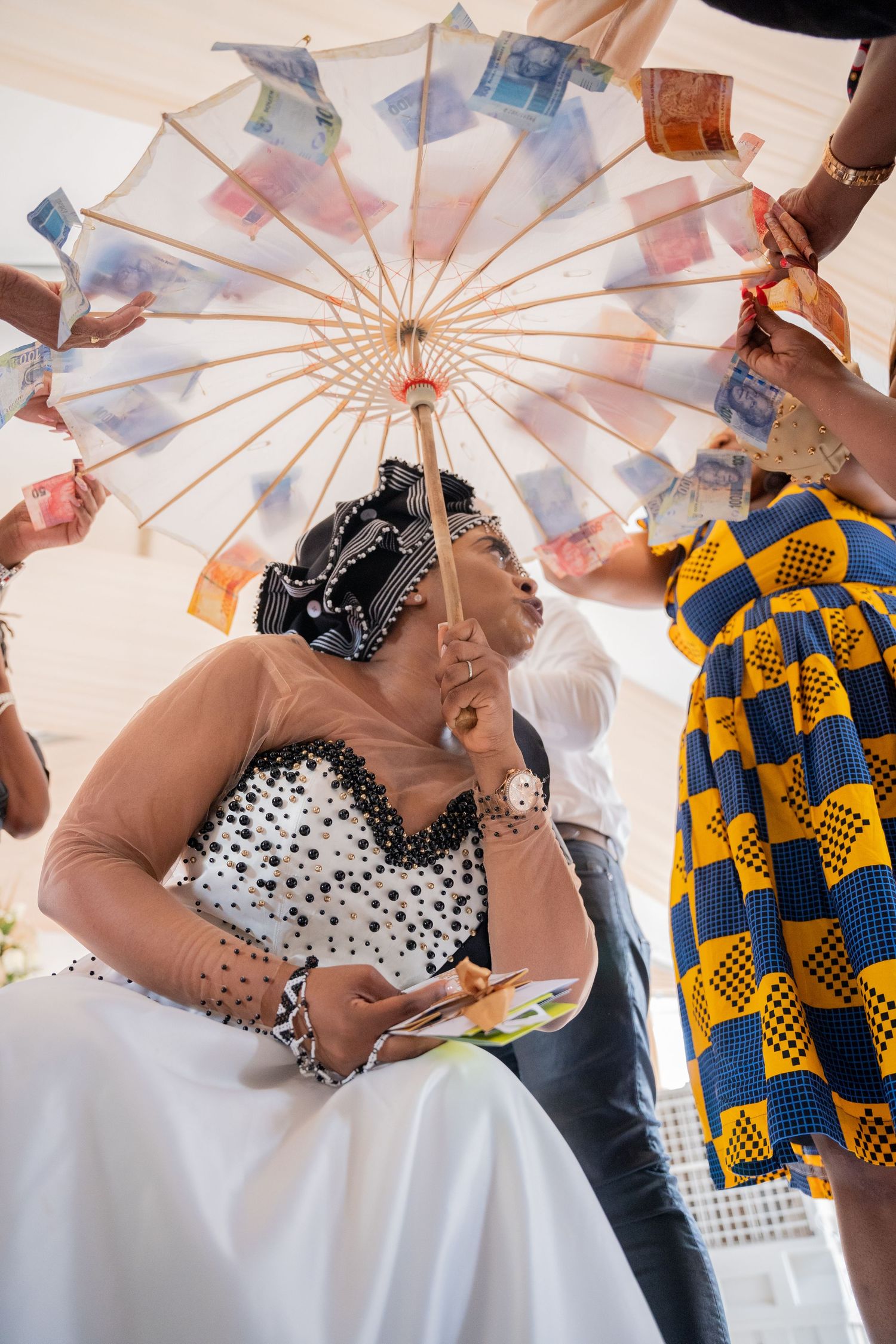 Bride in black and white beaded dress holding umbrella decorated with banknotes at African wedding.