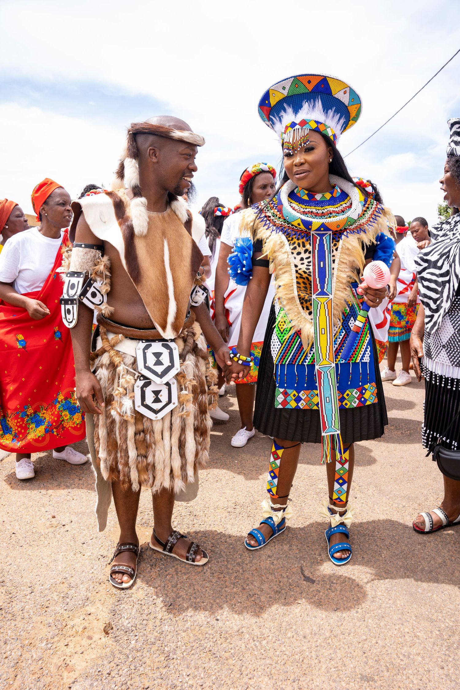 Zulu bride and groom in colorful traditional beaded attire and animal skin garments during outdoor wedding celebration.